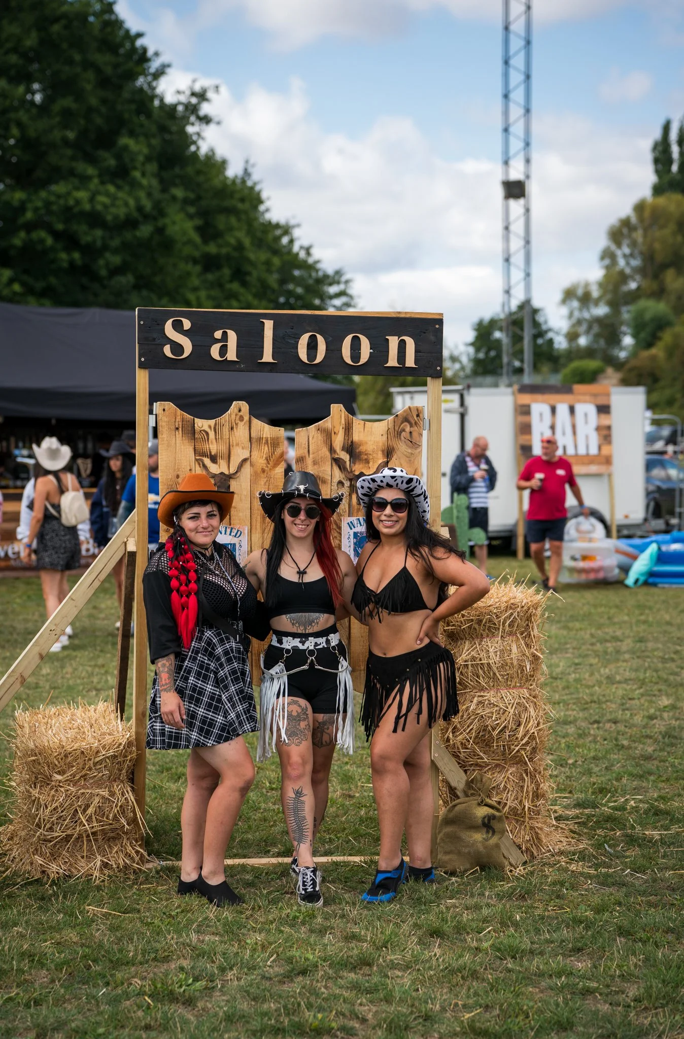 Three women dressed in cowgirl outfits standing in front of a saloon sign at an outdoor event. The women are smiling and wearing hats, sunglasses, and western-themed clothing, with hay bales and a wooden fence behind them.