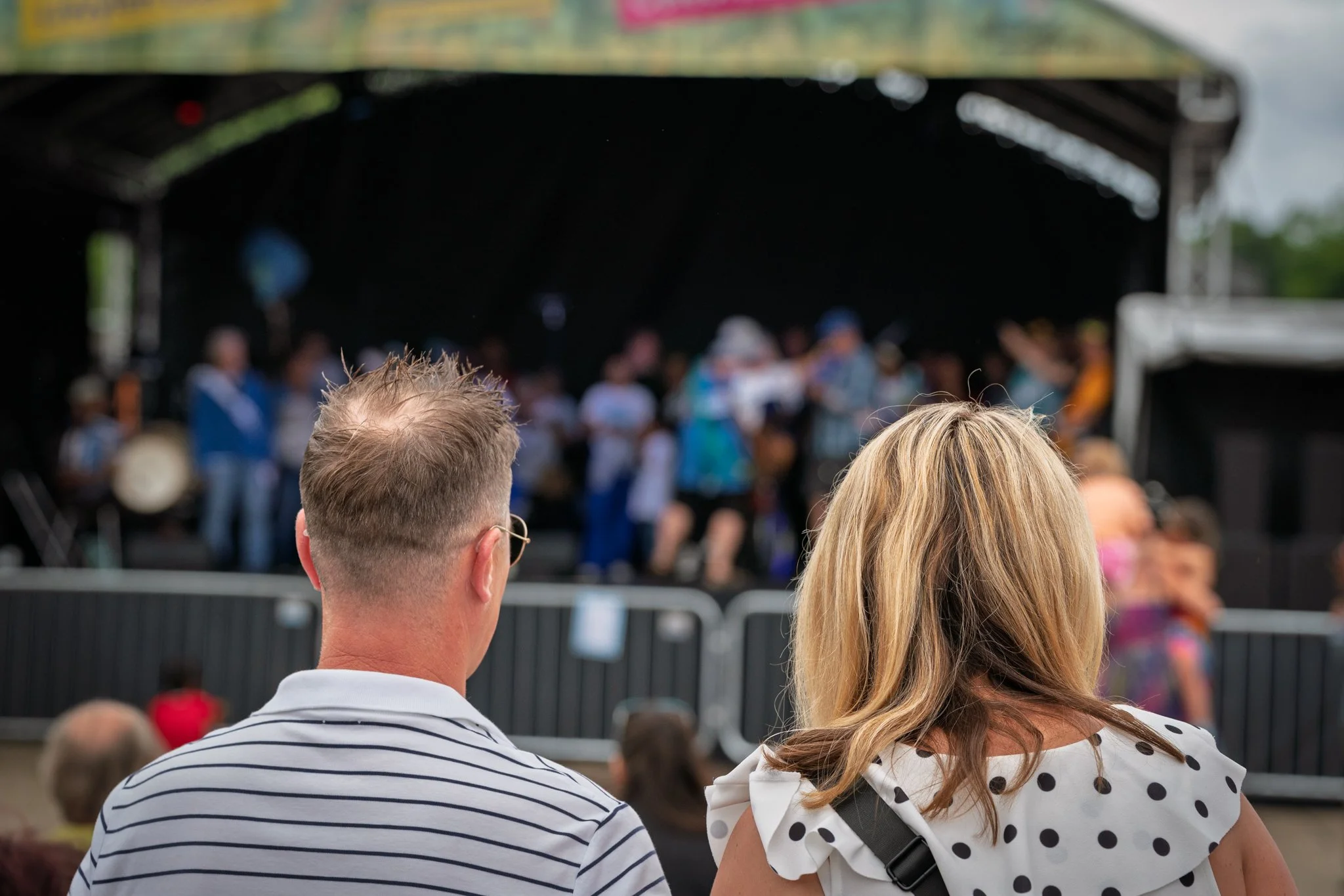 People watching a stage performance at an outdoor event, seen from behind.