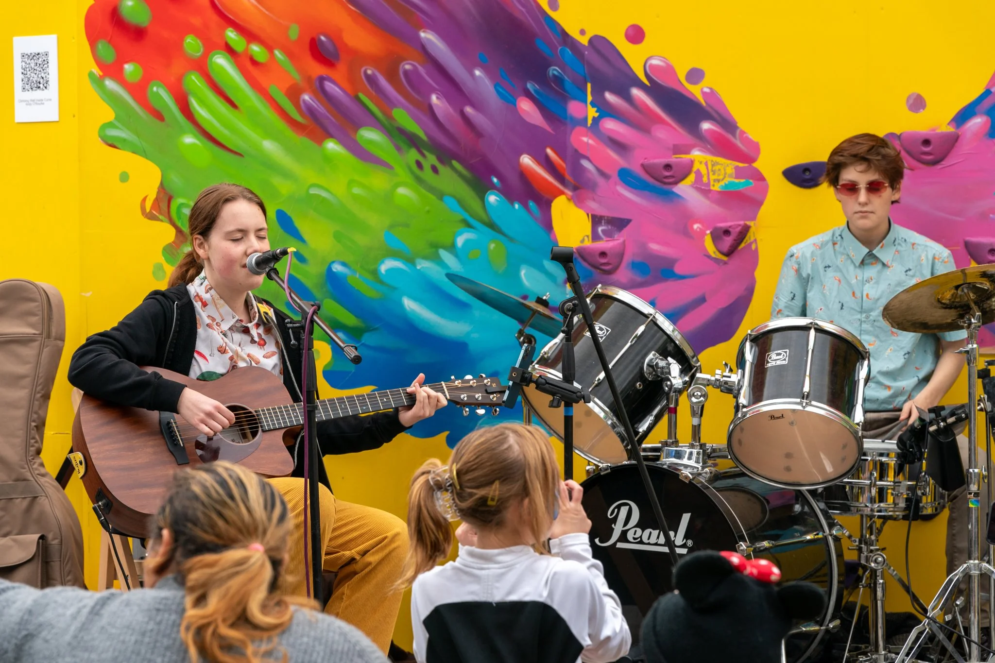 A young girl playing guitar and singing into a microphone, with a young boy standing behind a drum set, performing on a stage with a colorful graffiti-style mural in the background, and an audience of children watching.