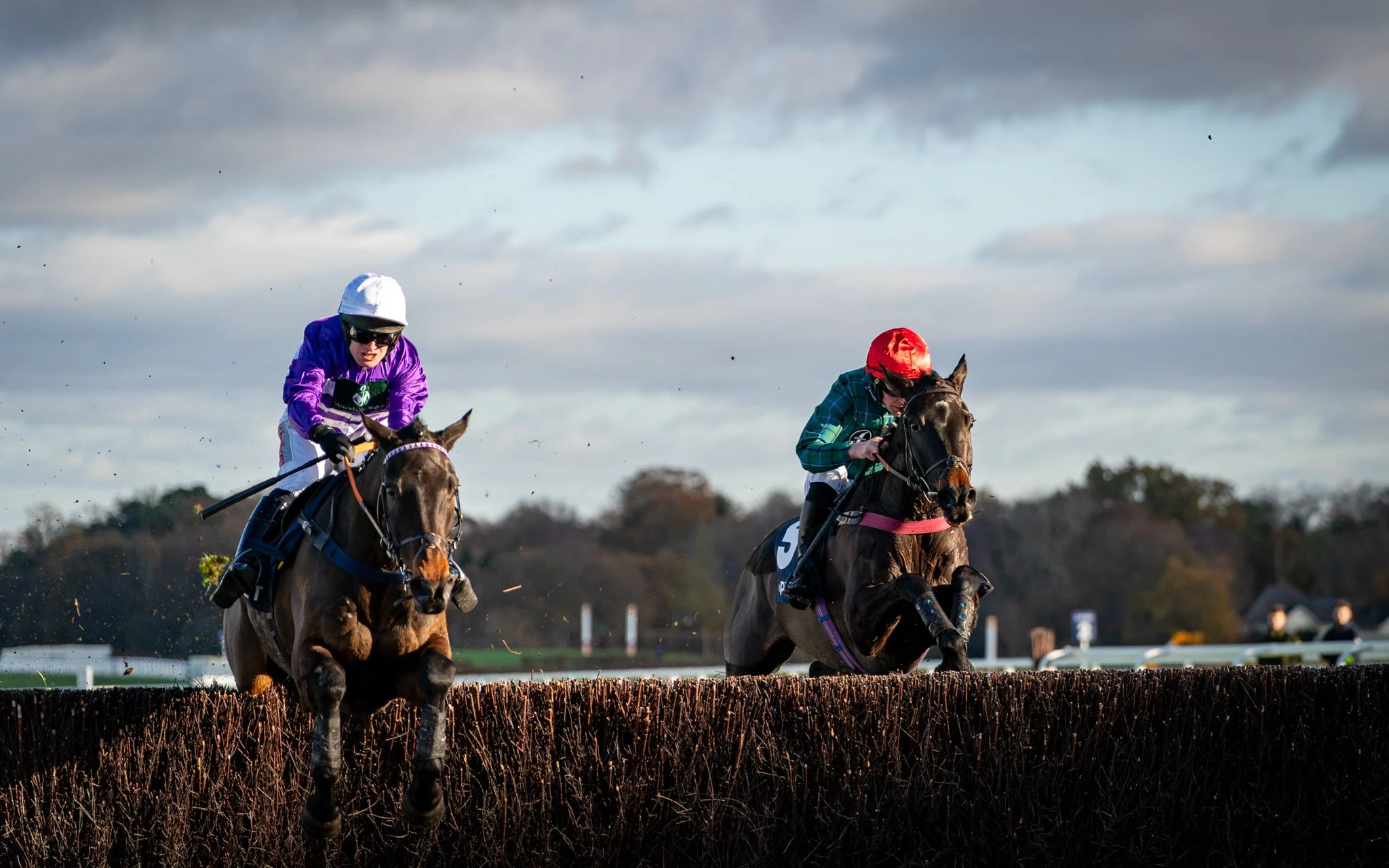 Jockeys riding horses in a race on a track outdoors, with a cloudy sky and trees in the background.