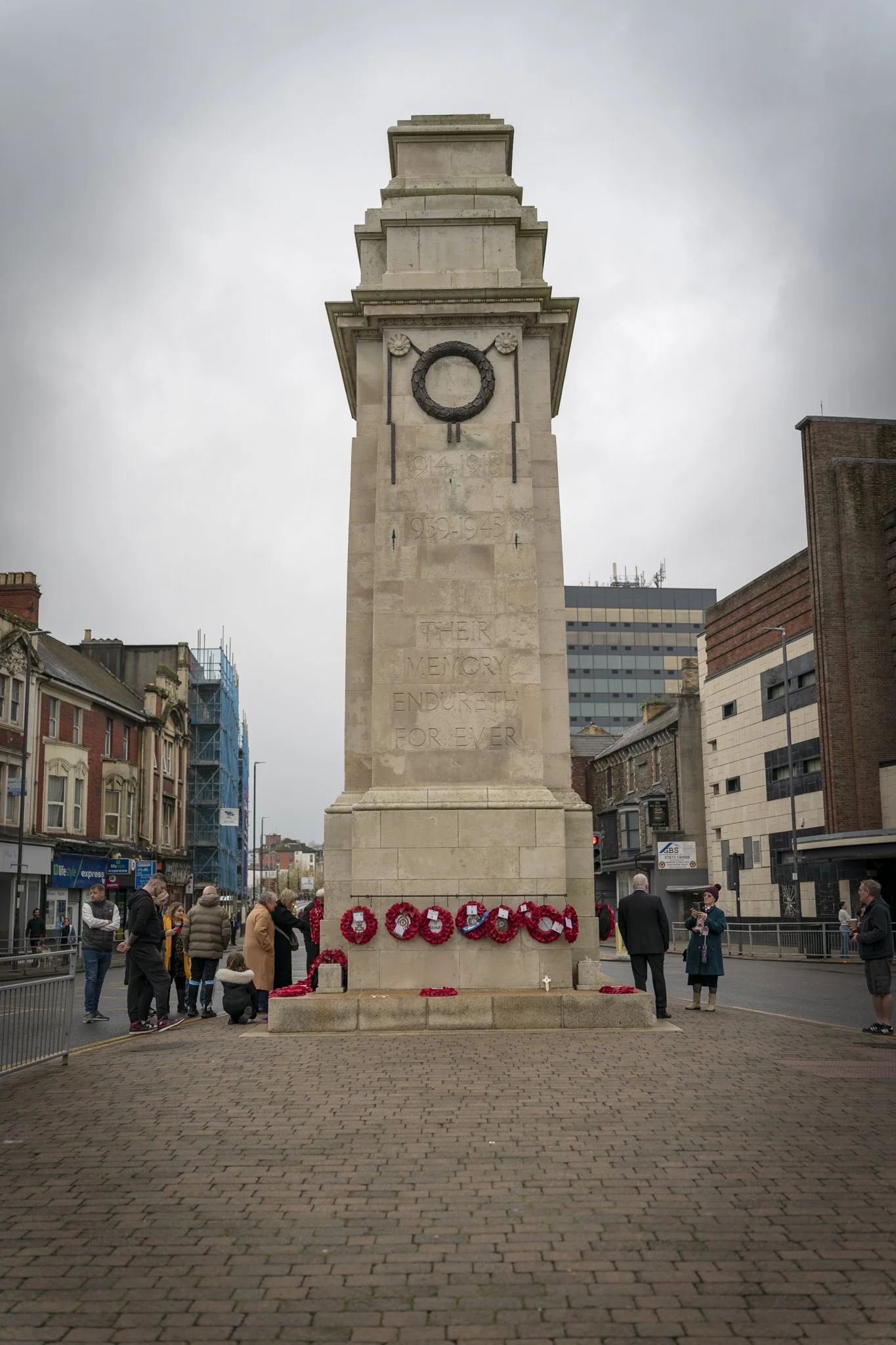 A war memorial with wreaths at its base in an urban setting with buildings and people around.