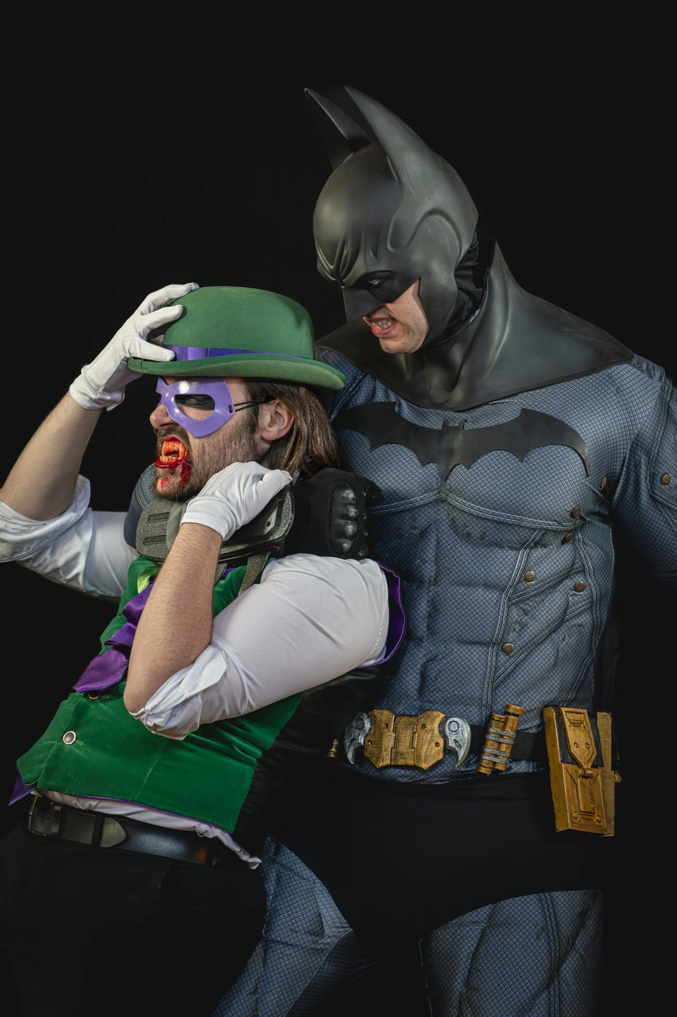 Costumed individuals dressed as Batman and the Joker, with Batman holding the Joker's hat and gripping him by the shoulder, set against a black background.
