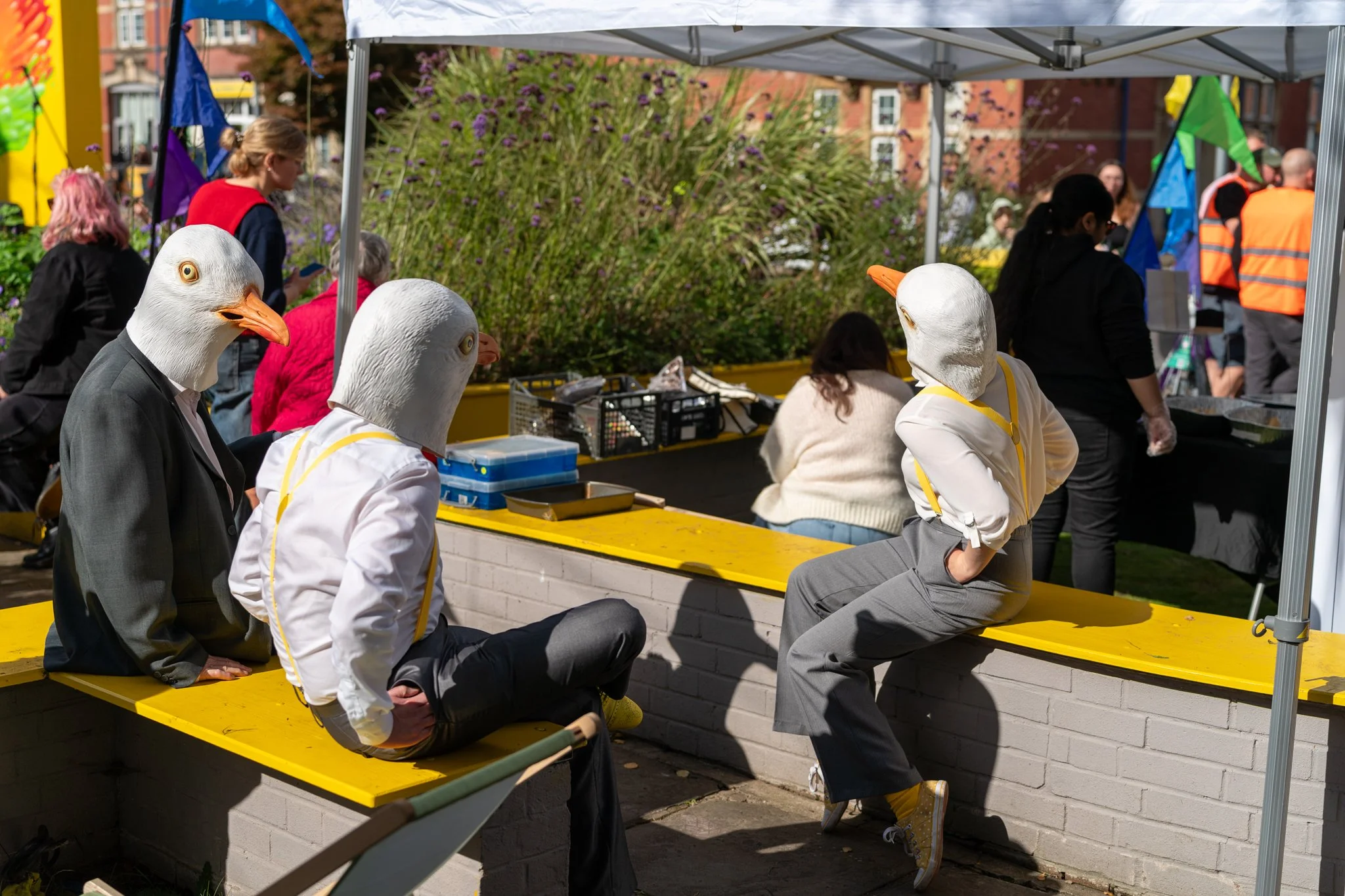 People at an outdoor market with some dressed in duck masks, sitting on a yellow bench under a white canopy.