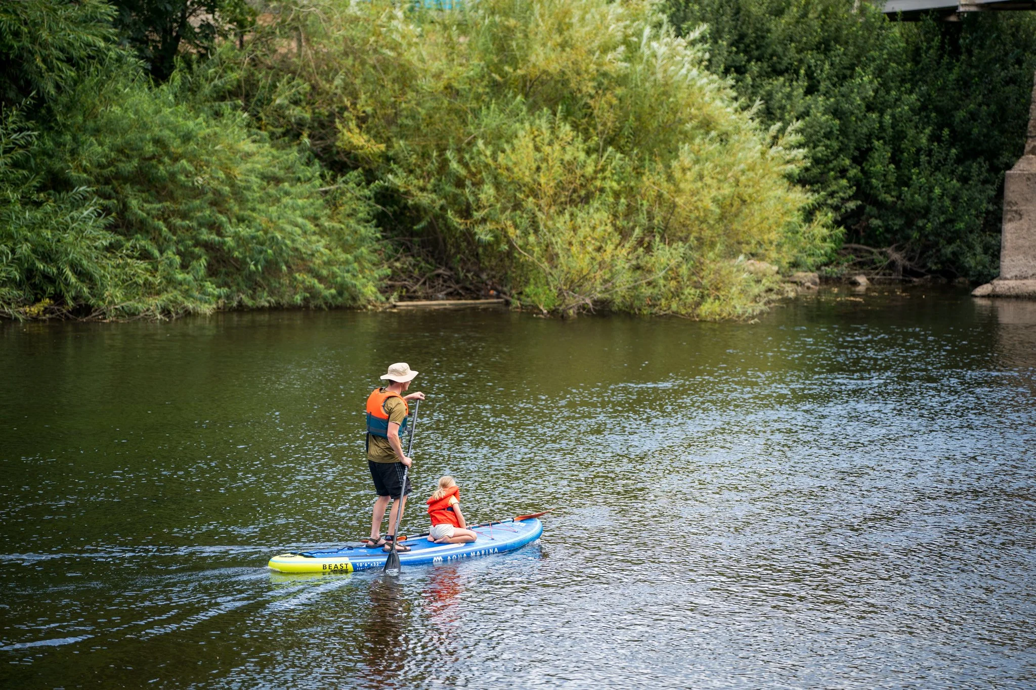 A person standing on a paddleboard with two children, one sitting and one kneeling, on a river surrounded by lush green trees.