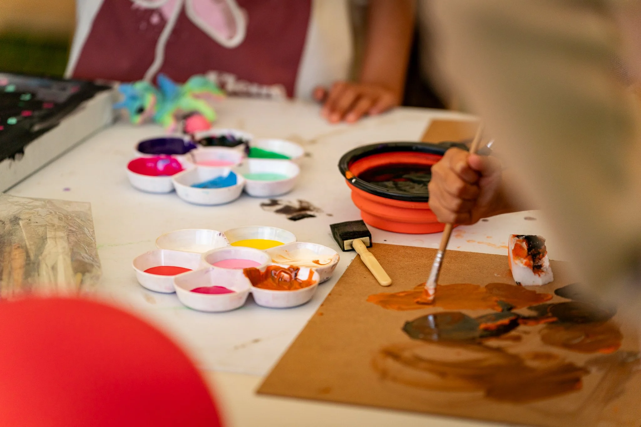 Child's hand holding a paintbrush while creating watercolor art with various paints on paper.