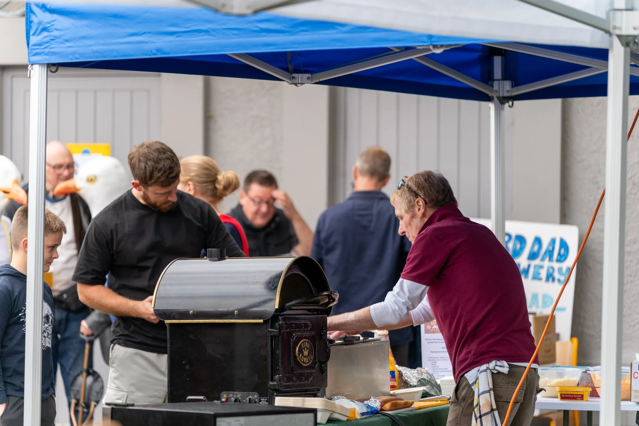 People gathered under a blue canopy at an outdoor event, with some individuals grilling food and others standing nearby.