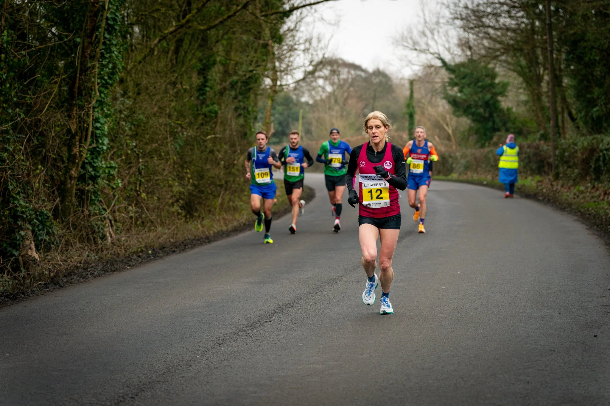 A group of runners participating in a race on a rural road surrounded by trees, with one woman in front wearing bib number 12, dressed in athletic gear, and other runners behind her wearing bibs numbered 109, 151, 79, and 86.