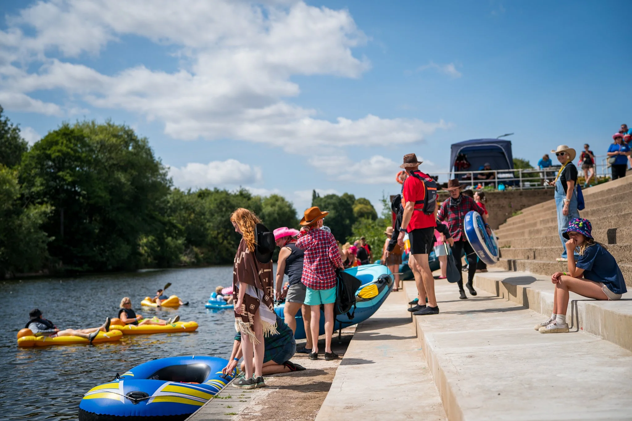 People gathered on a riverbank with kayaks and paddle boards, preparing to float on the water under a partly cloudy sky, with lush green trees in the background.