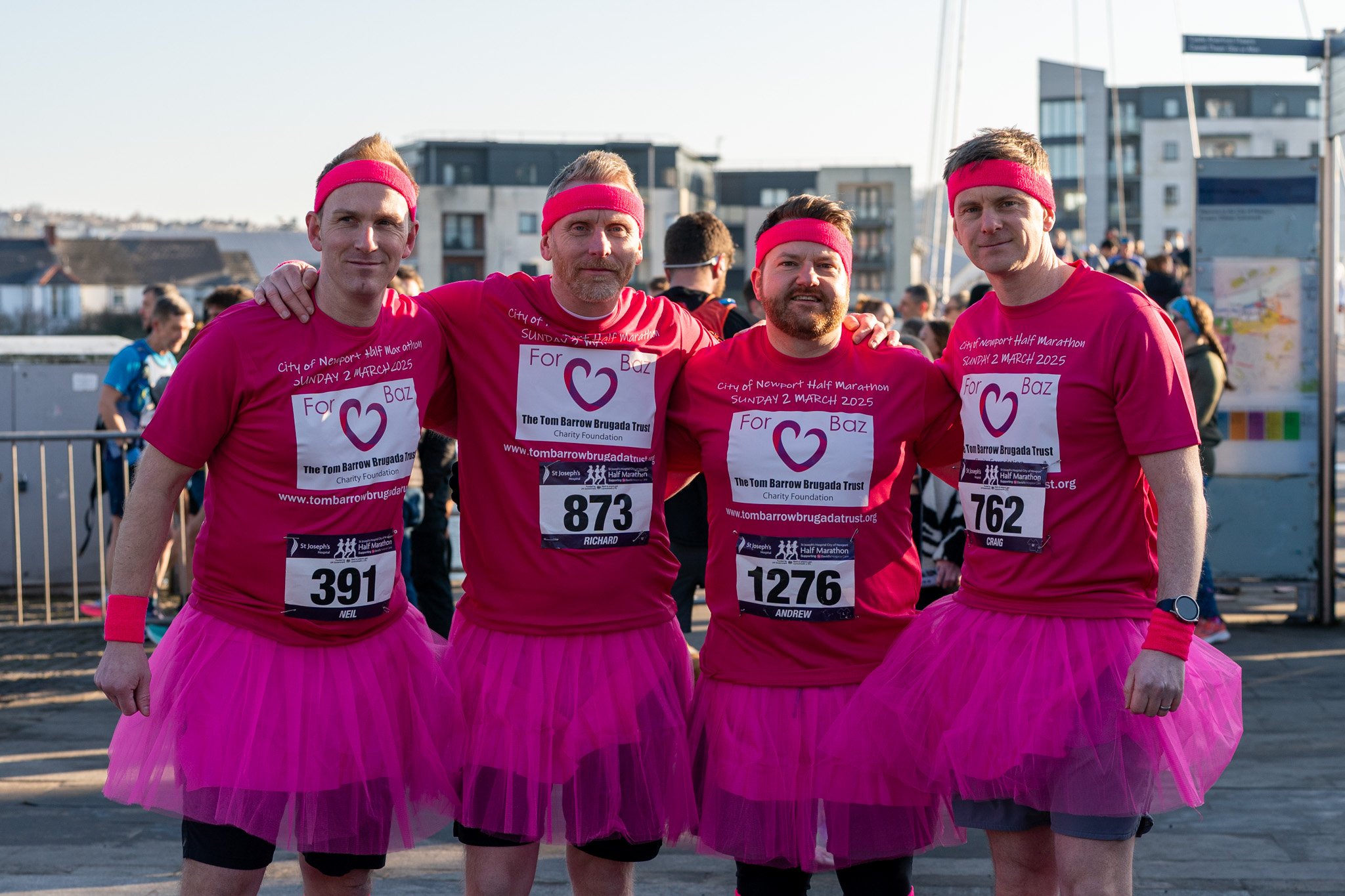 Four men dressed in bright pink T-shirts and pink tutus, wearing pink headbands, posing together at a marathon event with race bibs. They are smiling and standing outdoors with a crowd and buildings in the background, on a sunny day.