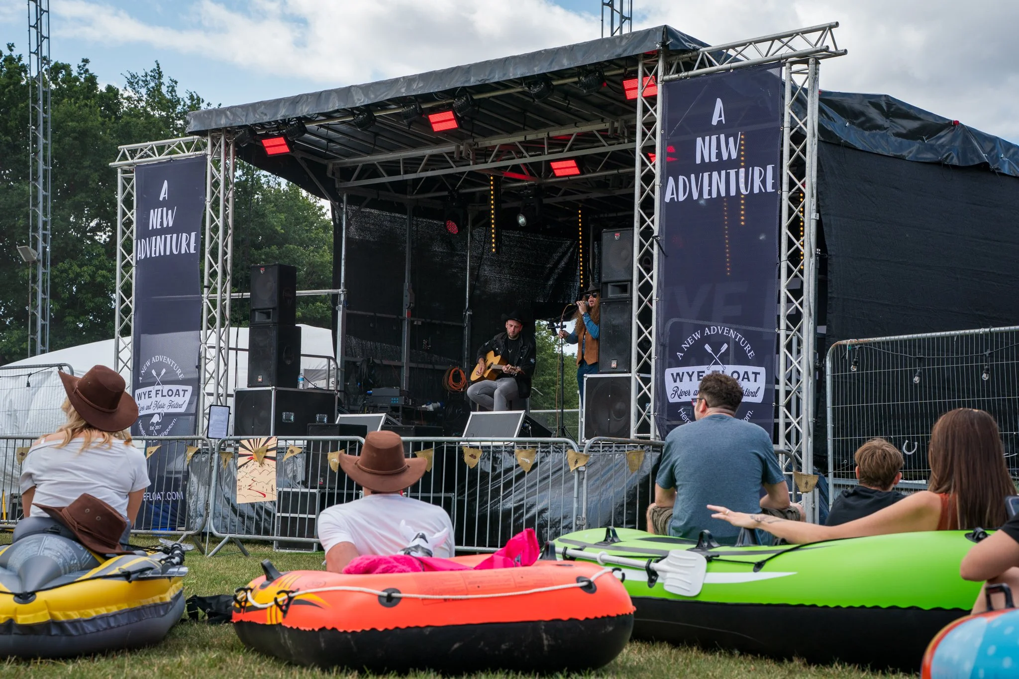 Outdoor music festival stage with performers and audience, kayaks and life jackets on grass in foreground, trees around, and cloudy sky.