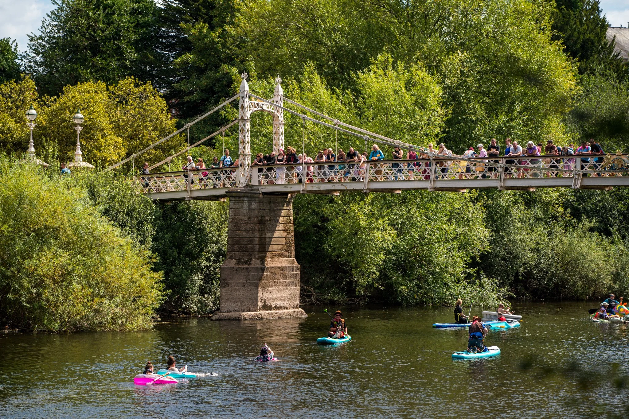 People paddleboarding on a river beneath a white suspension bridge with lush green trees in the background.