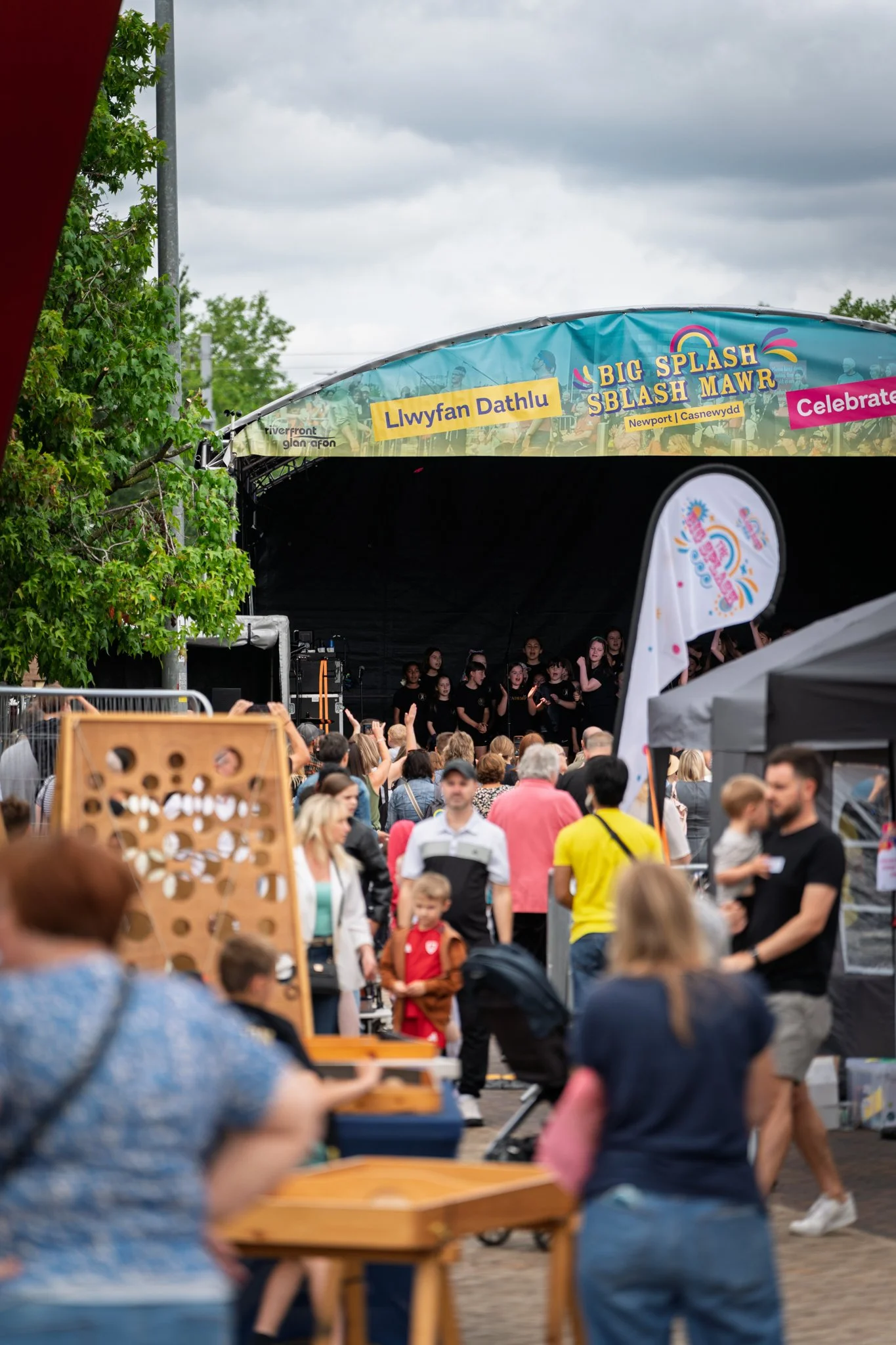 Crowd gathered at an outdoor event with a stage labeled 'Big Splash Sblash Mawr' and yellow banners with Welsh text, children and adults watching musical performers.