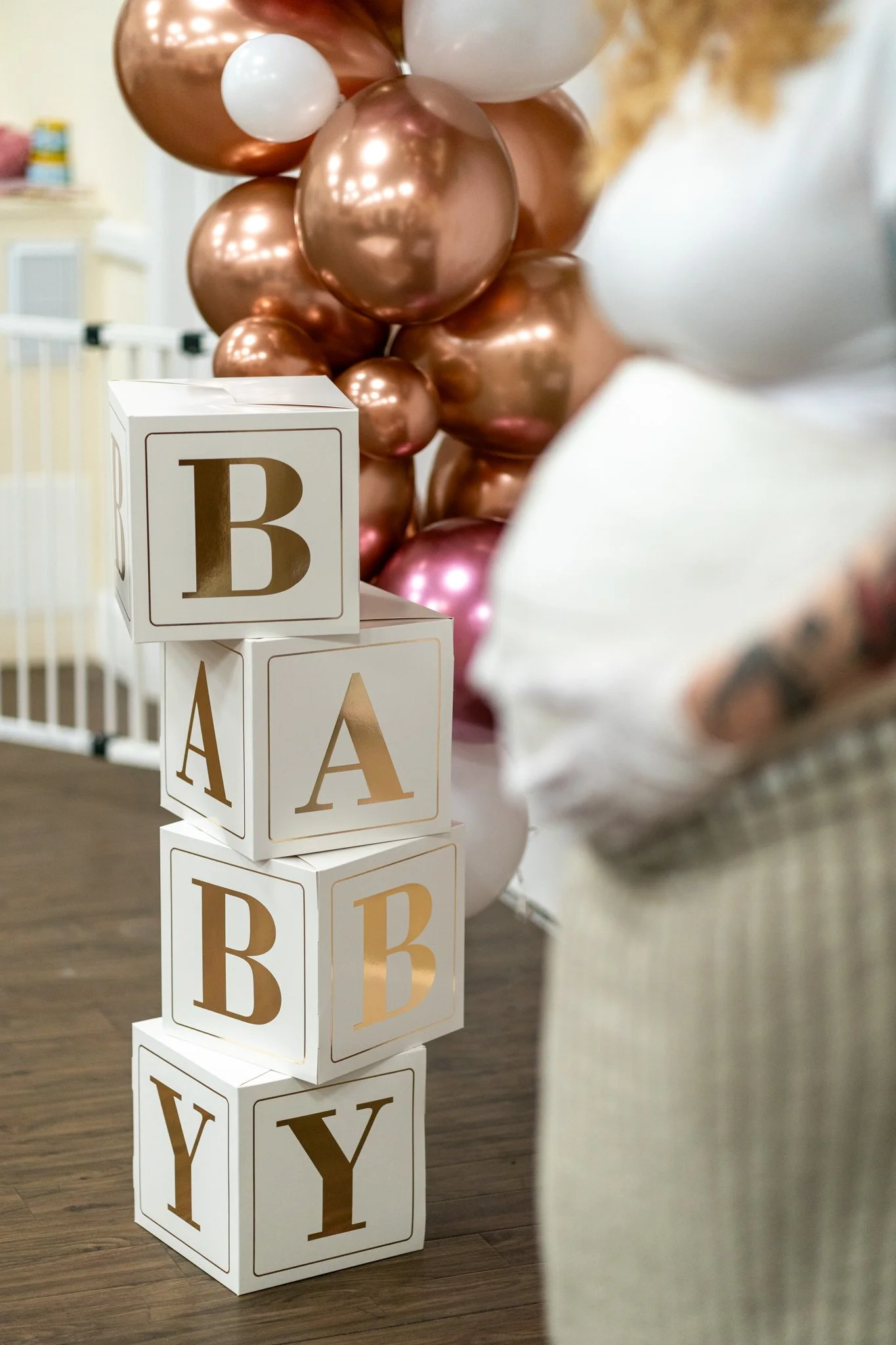 A tower of white blocks with gold and black alphabet letters, topped with a cluster of roses gold, white, and pink balloons, on a wooden floor in a room with a baby gate in the background.