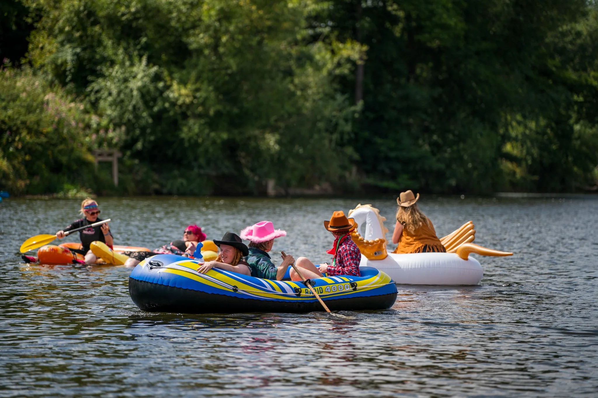 People in colorful and themed hats relaxing on inflatable boats and kayaks on a river, with a lush green forest in the background.