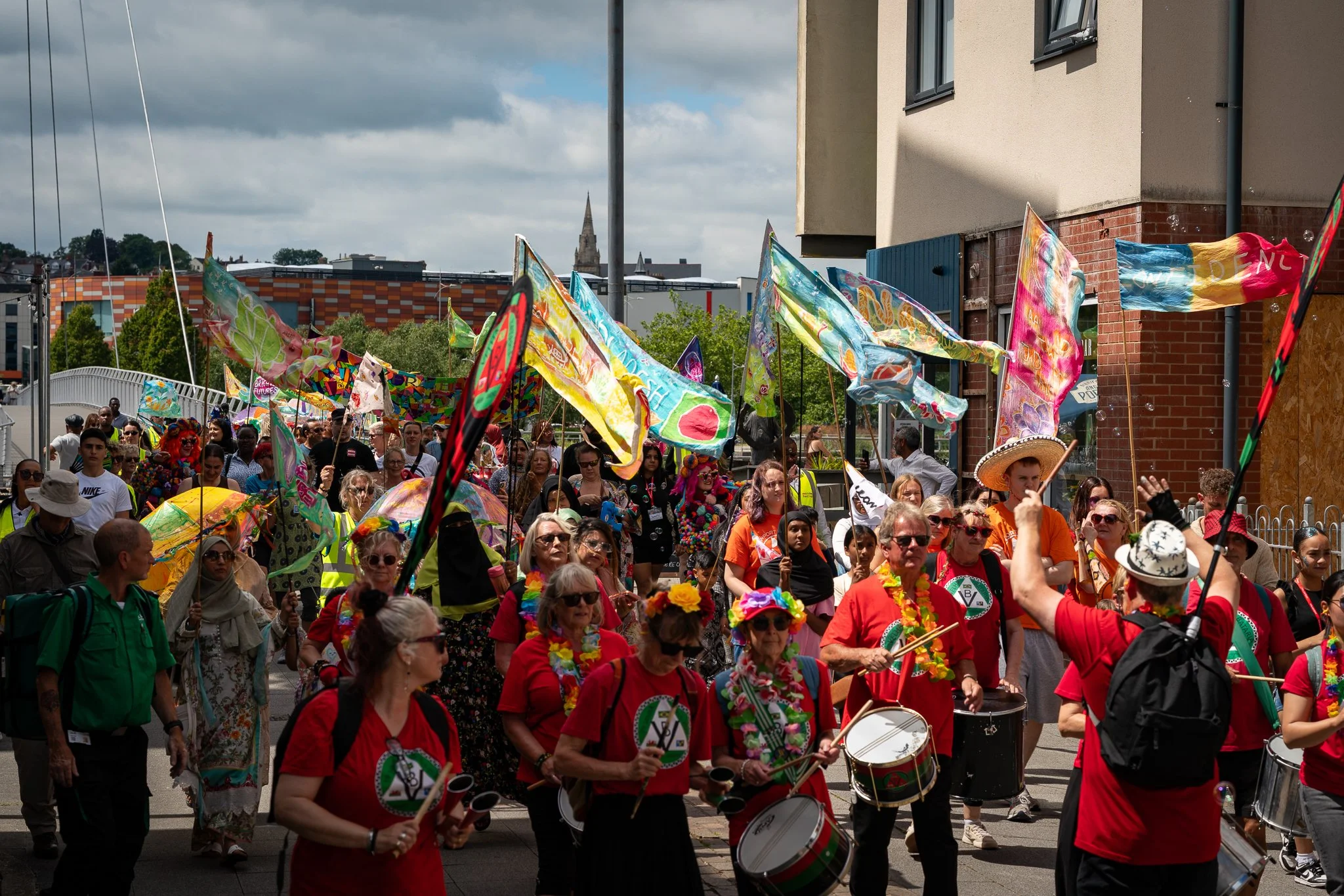 A crowd of people in colorful costumes and accessories participating in a parade or festival, waving flags, playing drums, and enjoying the outdoor event.