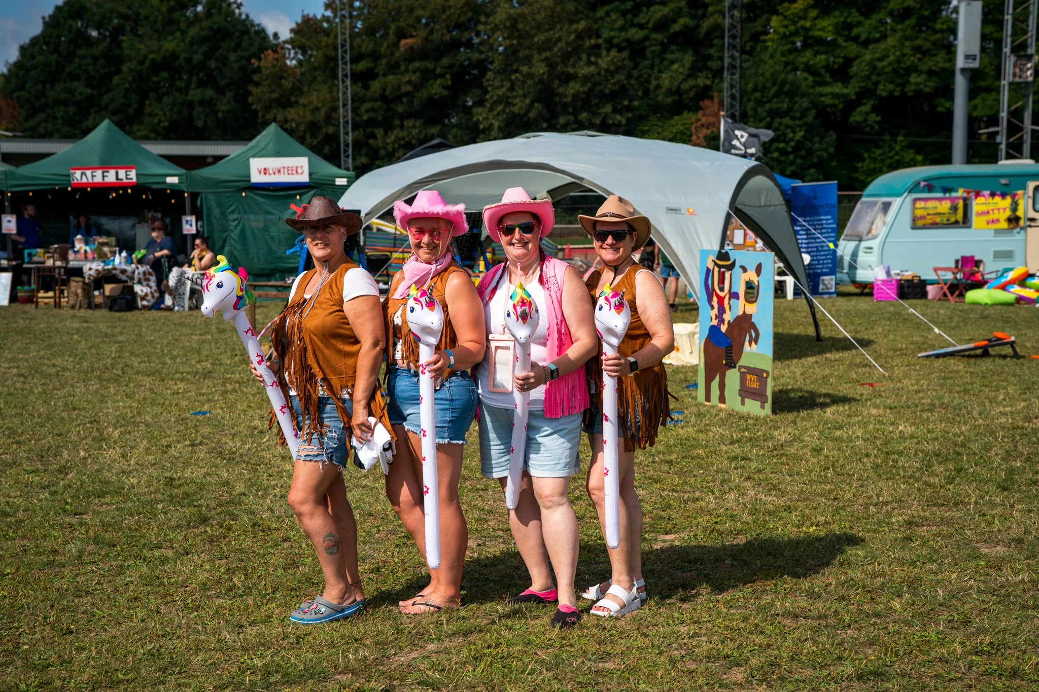 Four women dressed in cowboy costumes with inflatable unicorns, standing outdoors at a fair or festival, with tents and trees in the background.