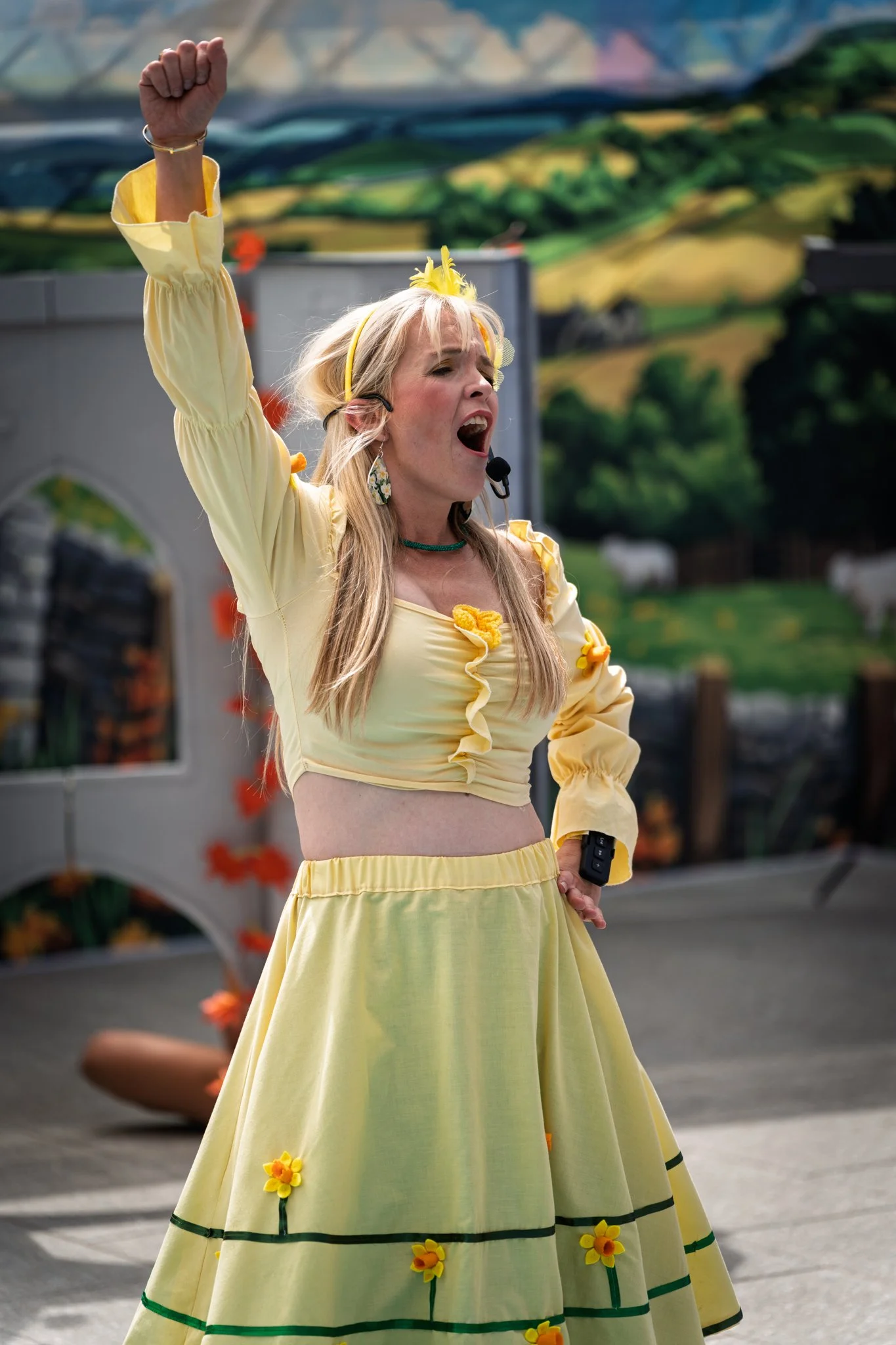 A woman wearing a yellow dress with flower decorations and a yellow headband, raising her fist in the air, appears to be singing or shouting during an outdoor event with a colorful landscape mural in the background.