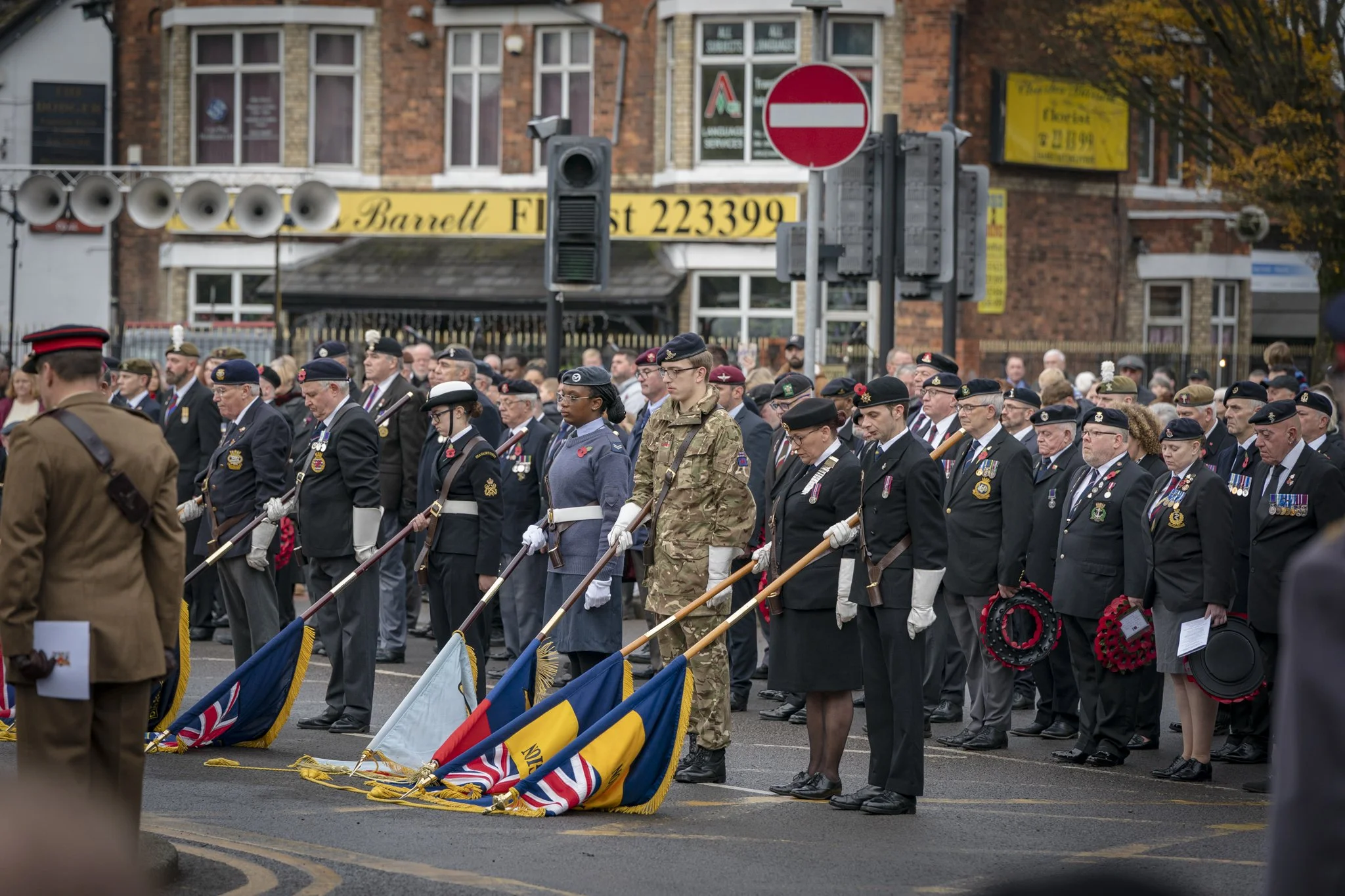 A large group of people, including military personnel and civilians, participating in a memorial or remembrance event on the street in front of brick buildings. They are standing with flags and wearing medals and poppies, paying respects.