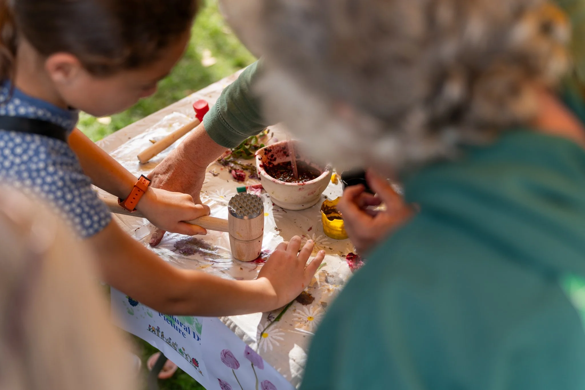 Children and adults engaged in outdoor crafting or art activity around a table covered with a floral tablecloth, using various tools and materials.