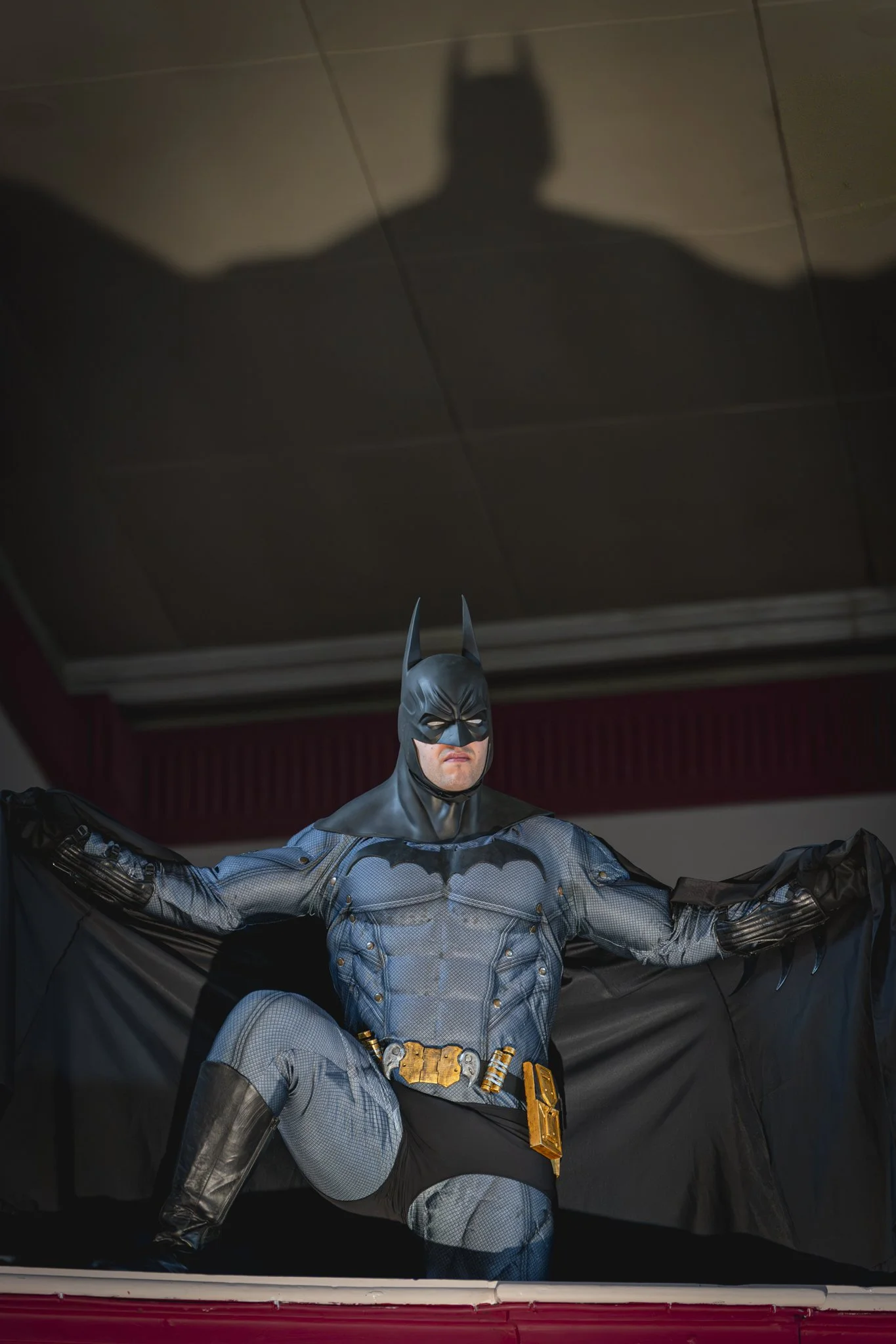 A person dressed as Batman standing with arms extended, wearing a blue and gray suit with a black cape and mask, with a shadow of Batman cast on the ceiling above.