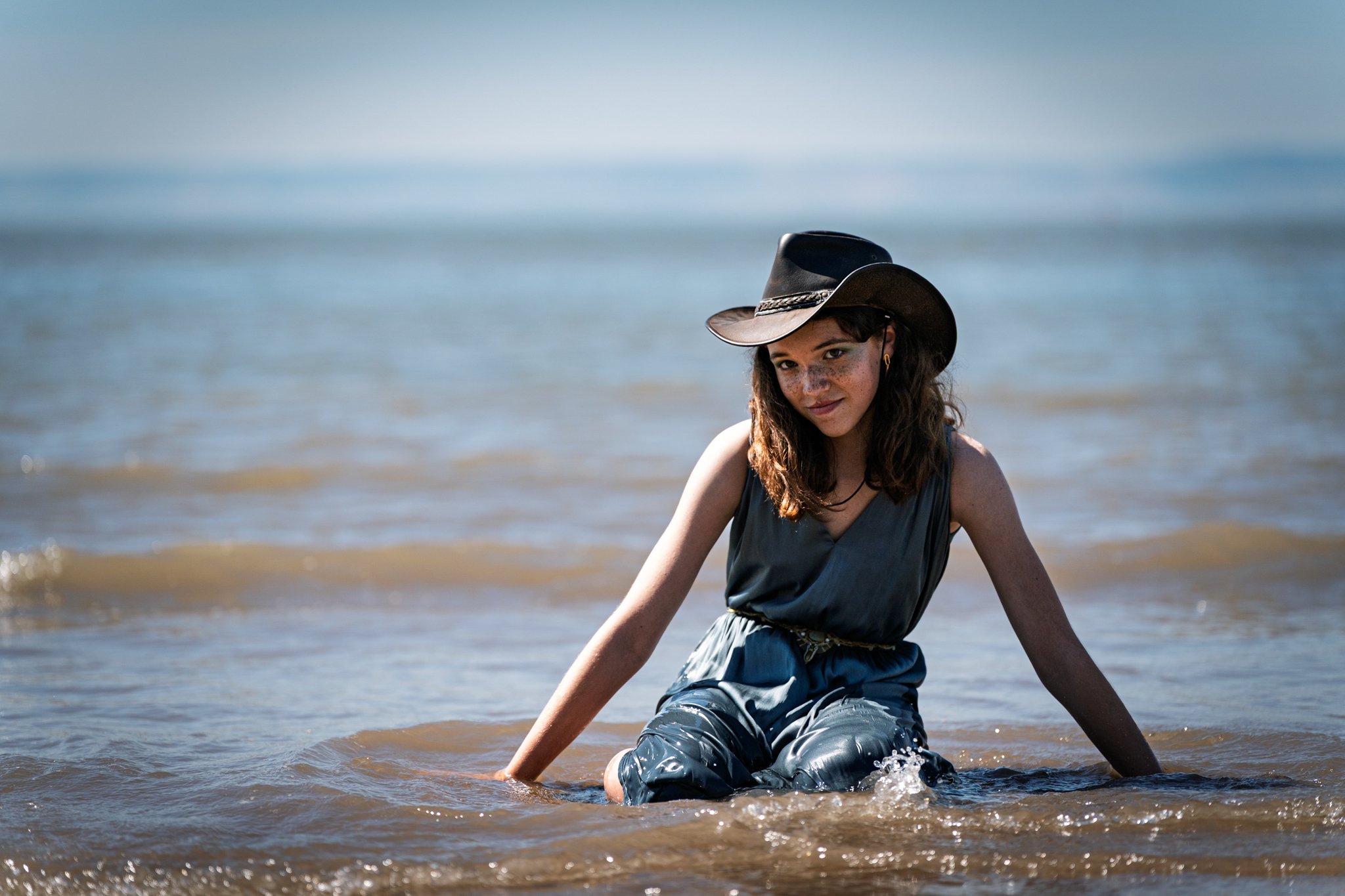 A woman with a cowboy hat sitting in shallow ocean water at the beach, smiling softly, wearing a dark sleeveless top and patterned pants.