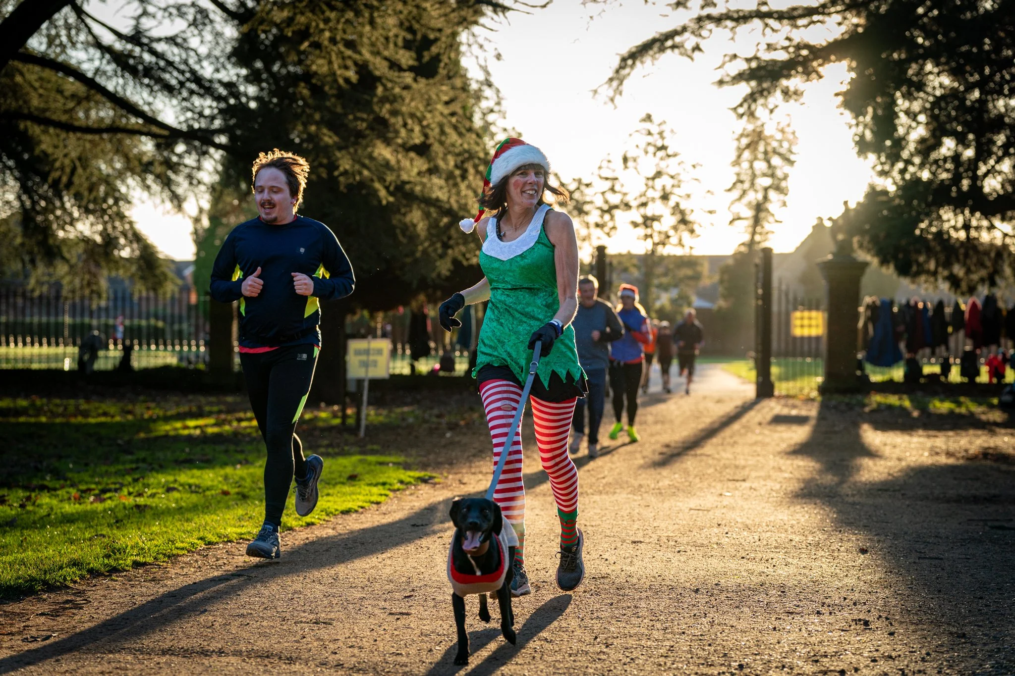 A woman dressed as an elf with red and white striped leggings, a green dress, and a Santa hat walking her dog in a park during sunset; a man jogging nearby, both surrounded by tall trees and other park visitors.