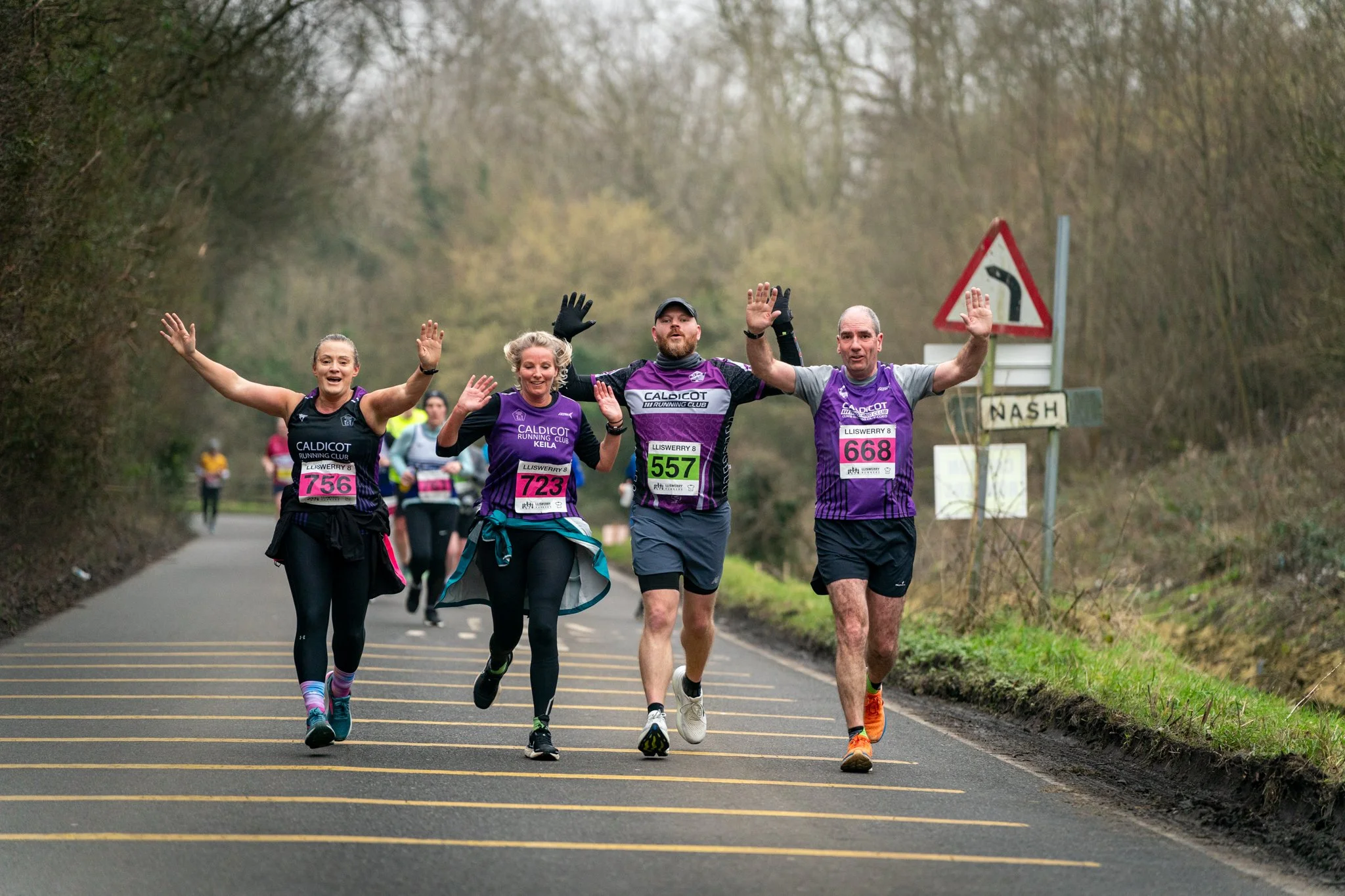 Group of marathon runners running on a rural road surrounded by trees, waving their hands at the camera.