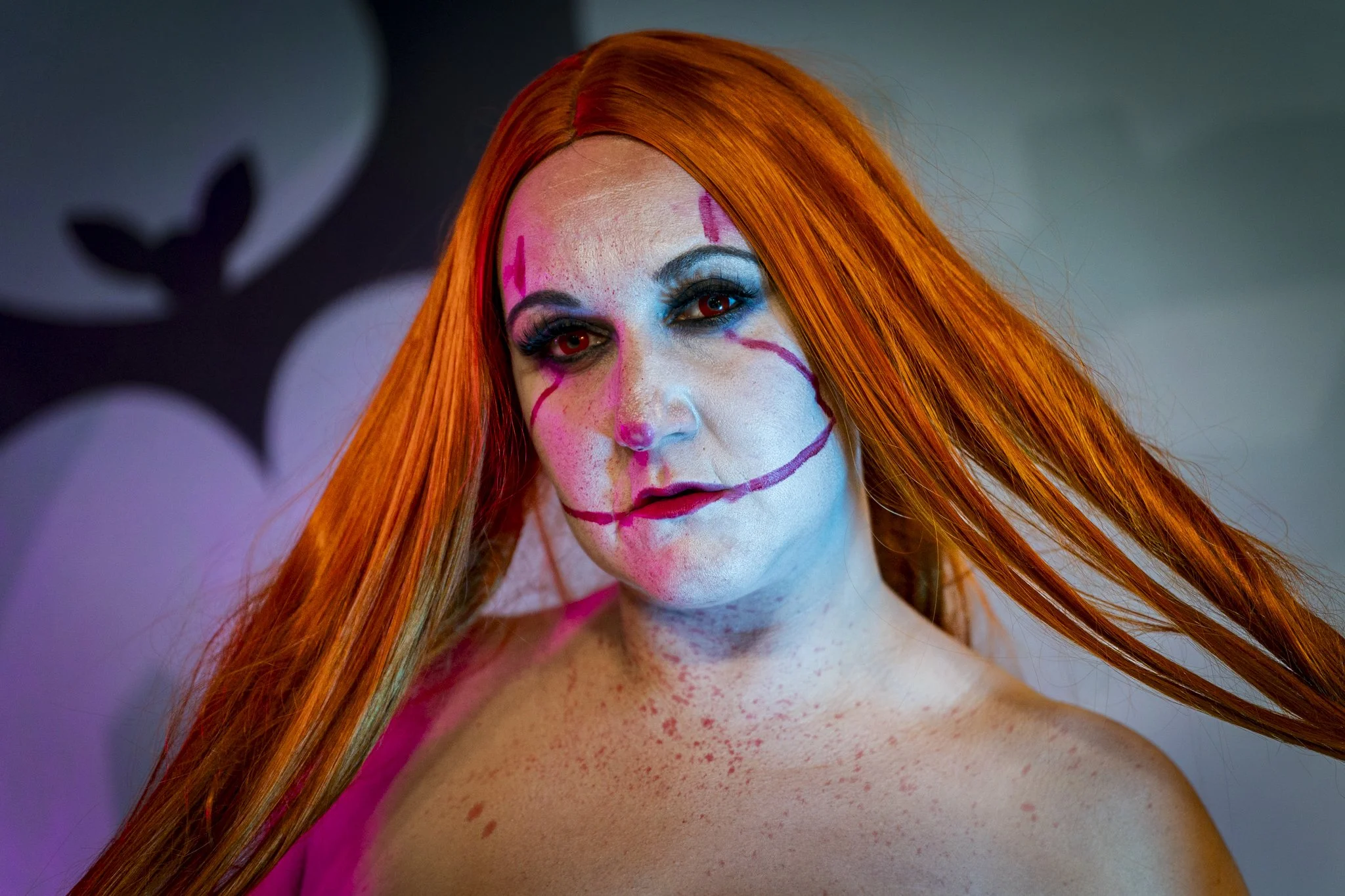 A woman with bright orange hair and clown makeup, including white face paint, pink and blue eye makeup, red lipstick, and painted pink lines on her face and chest.
