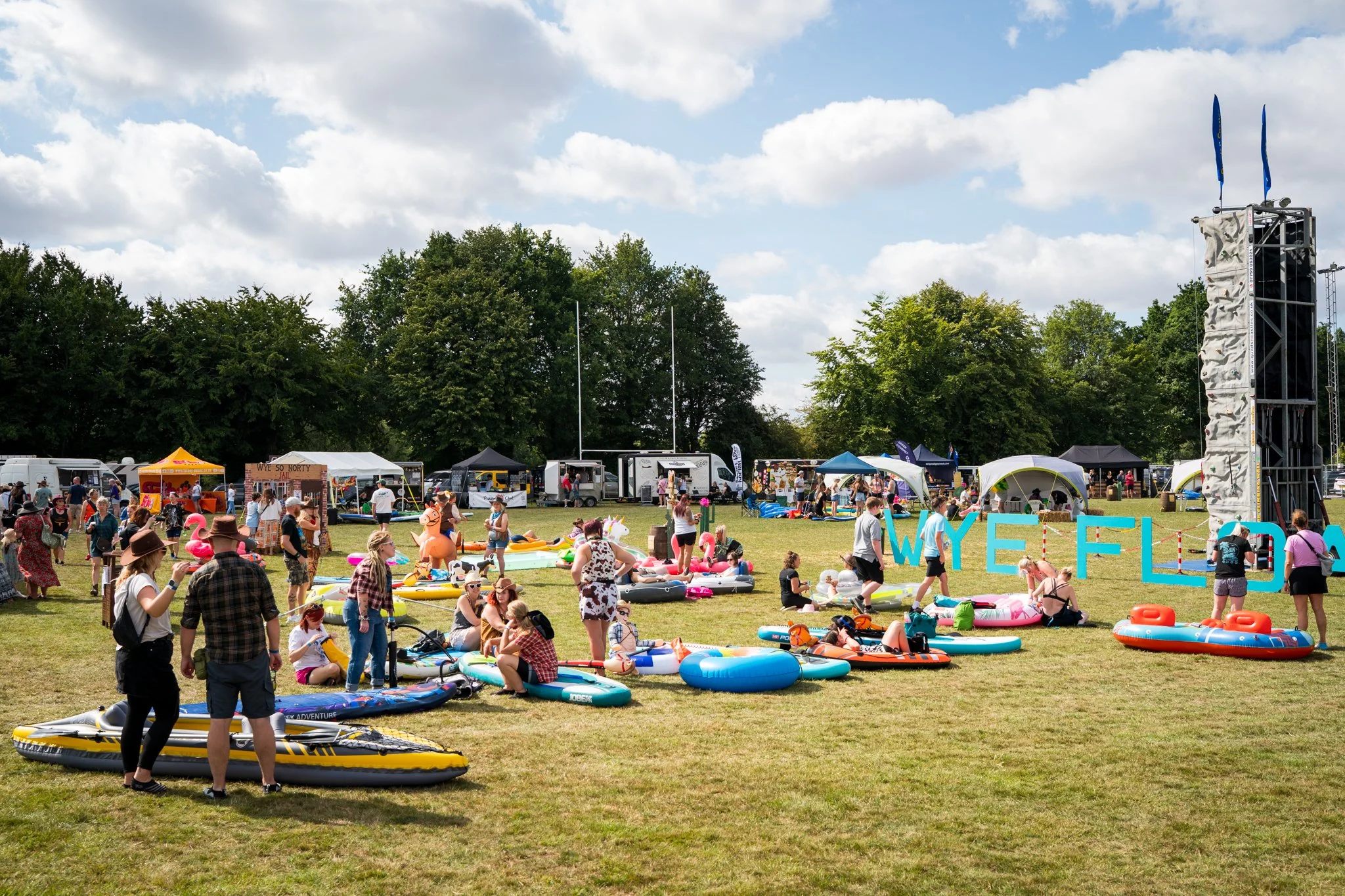 Crowd at a summer outdoor festival with inflatable pools, kayaks, and tents, under a partly cloudy sky.