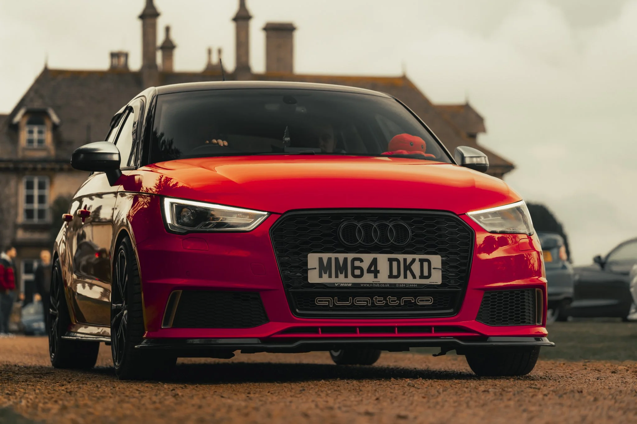 Red Audi Quattro car with UK license plate parked on a dirt ground in front of a historic building, with people in the background.