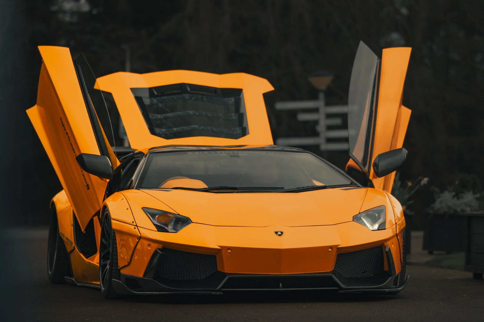 Front view of a bright orange Lamborghini sports car with scissor doors open, parked outdoors at dusk or nighttime.