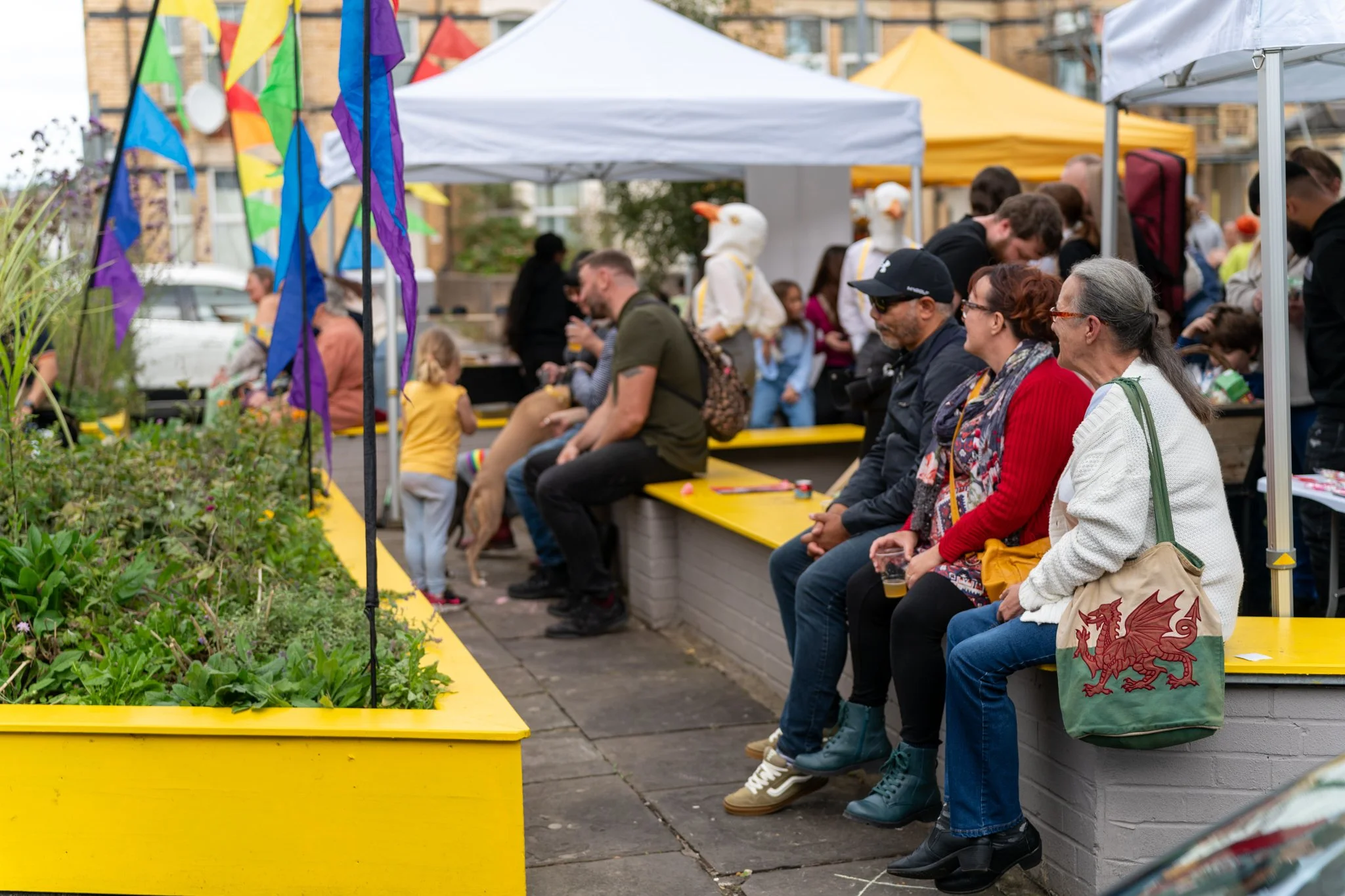 People sitting on a yellow bench at an outdoor event with colorful flags, tents, and a person dressed as a goose in the background.
