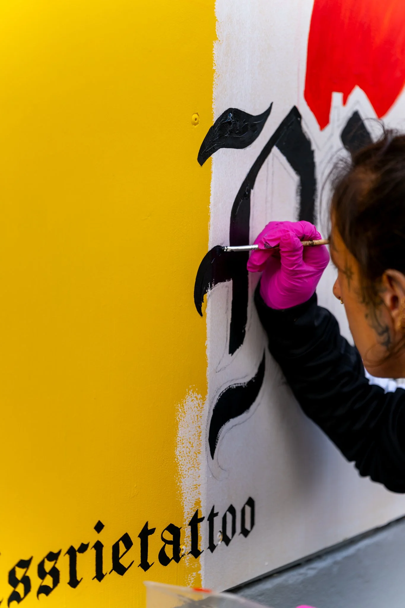 Person painting a mural with black and red paint on a white wall, wearing pink gloves.