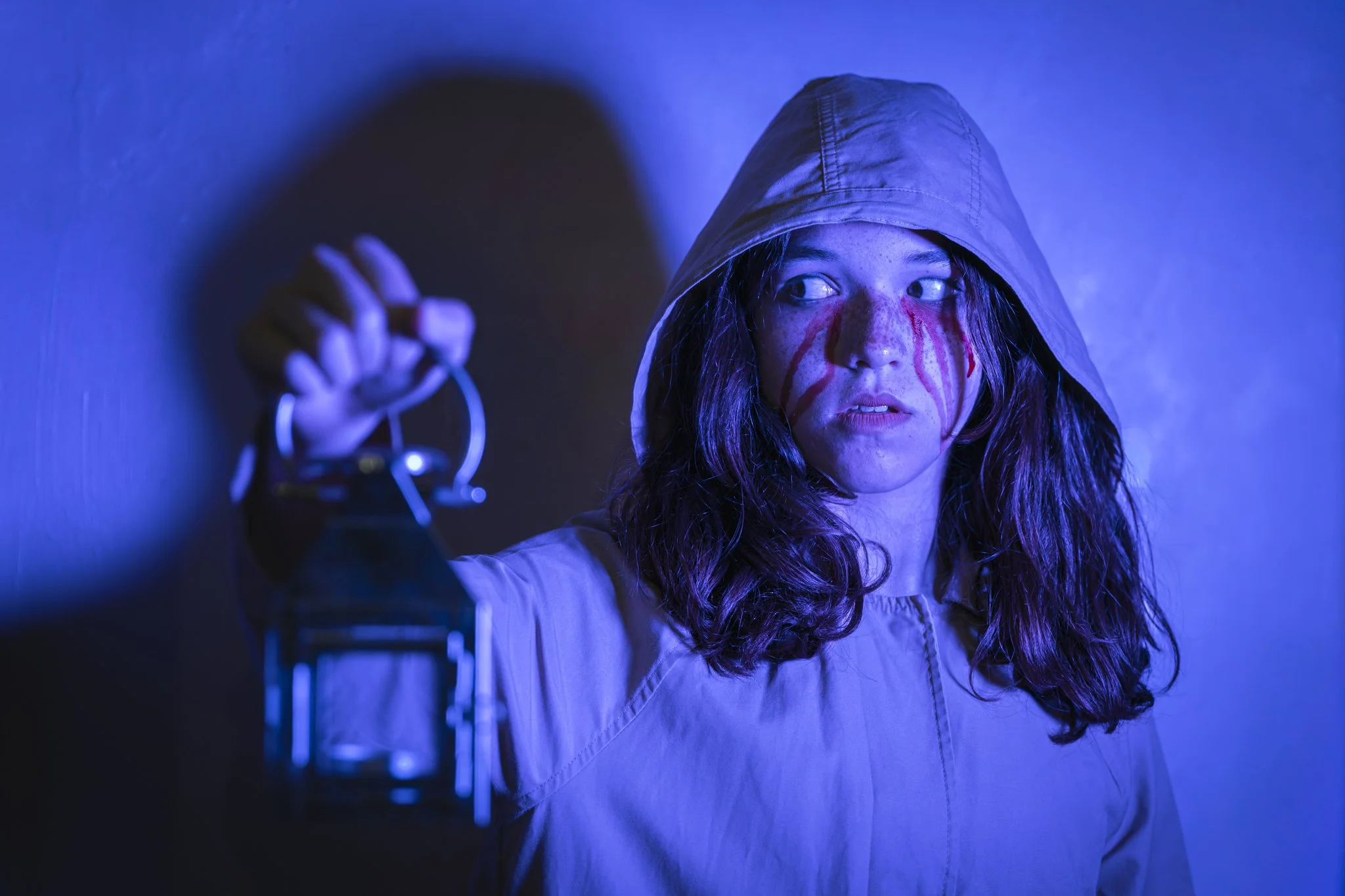 A young girl with dark hair, wearing a hooded jacket, is looking to the side with a serious expression. She has blood on her face and is holding spray bottles, with a dark background illuminated by blue light.