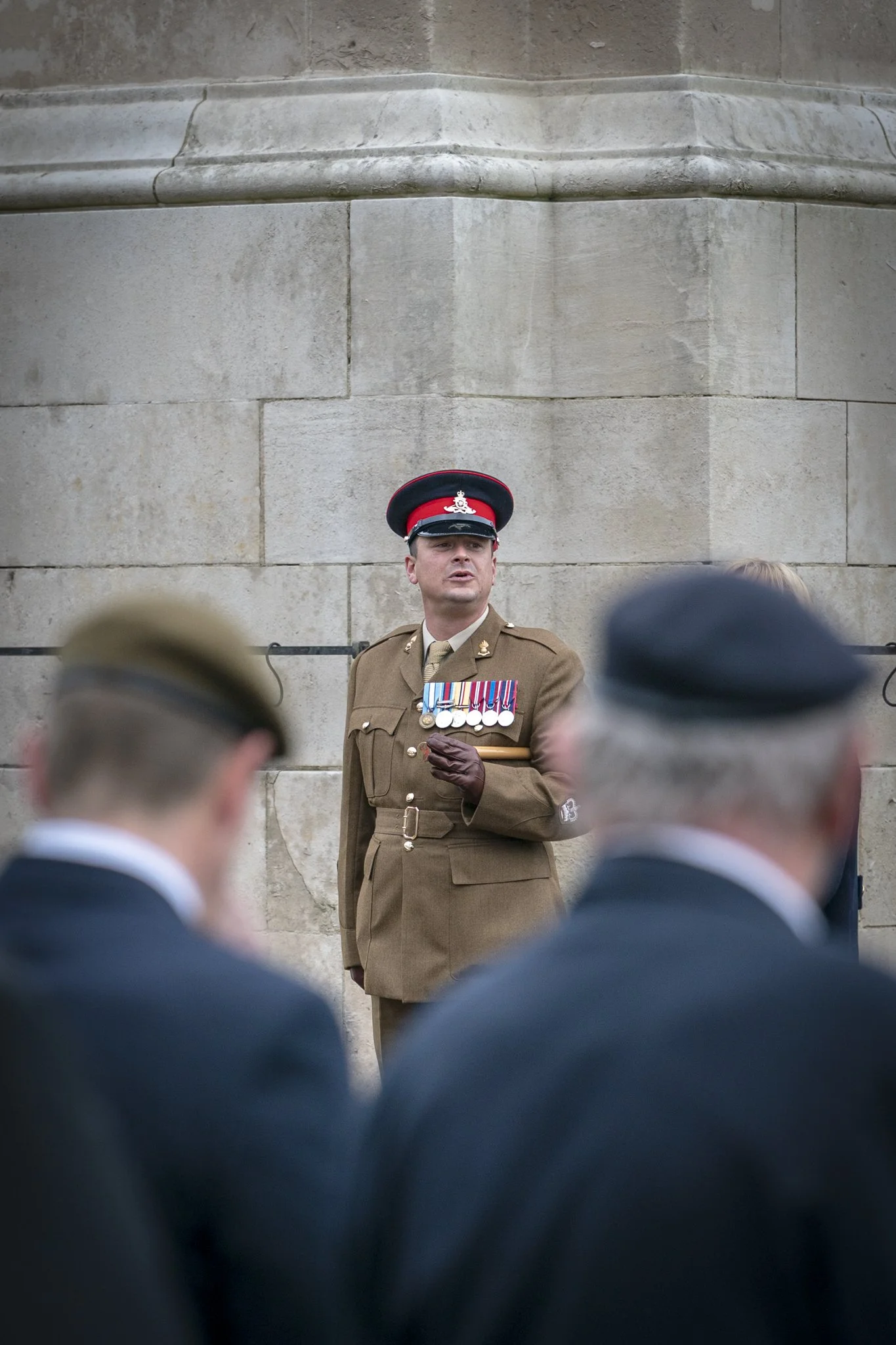 A military officer in uniform with medals is speaking at a ceremony, standing against a stone wall. The foreground shows blurred heads of other uniformed individuals.
