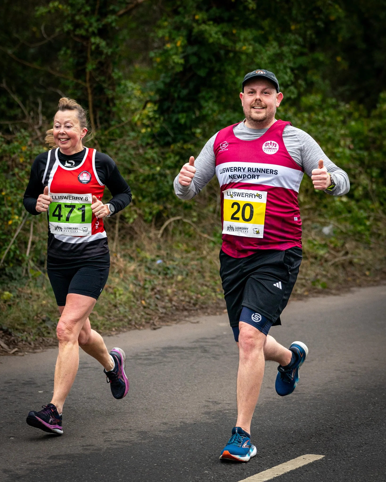Two runners participating in a race or marathon, wearing race bibs, running on a paved path with trees in the background.