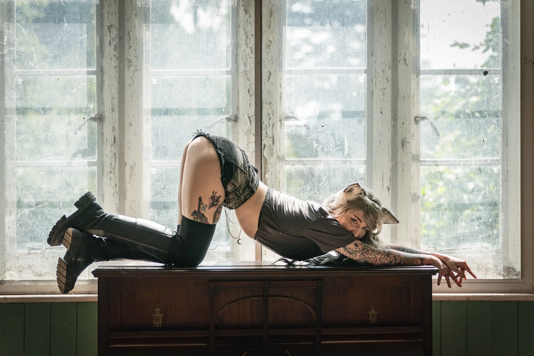 A woman with tattoos and dyed hair posing on a vintage piano, leaning forward with her arms extended, in front of large worn windows with peeling paint and a view of trees outside.