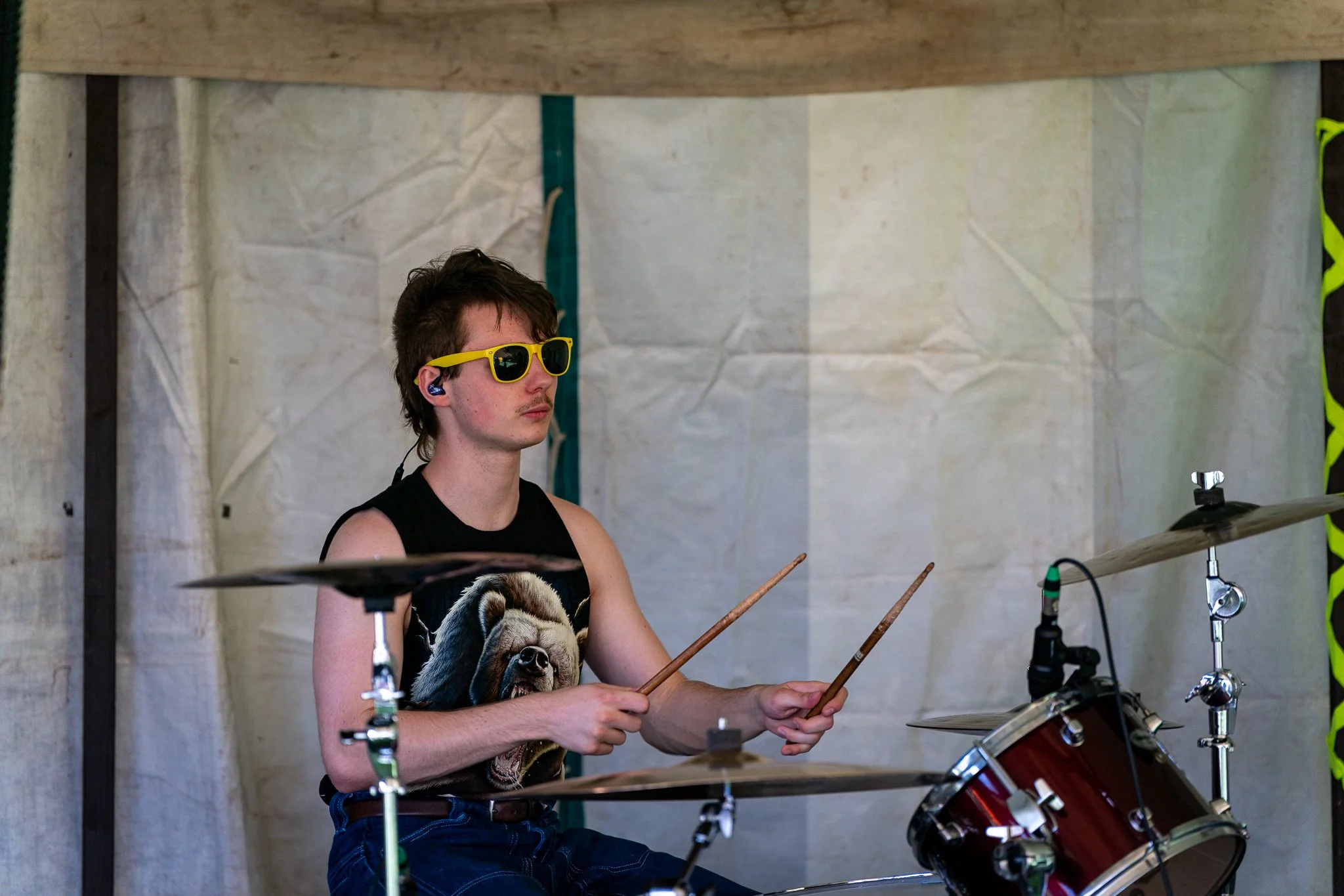 A young man wearing yellow sunglasses and a sleeveless shirt with a dog graphic is playing drums at an outdoor event, with a drum set and a tent in the background.