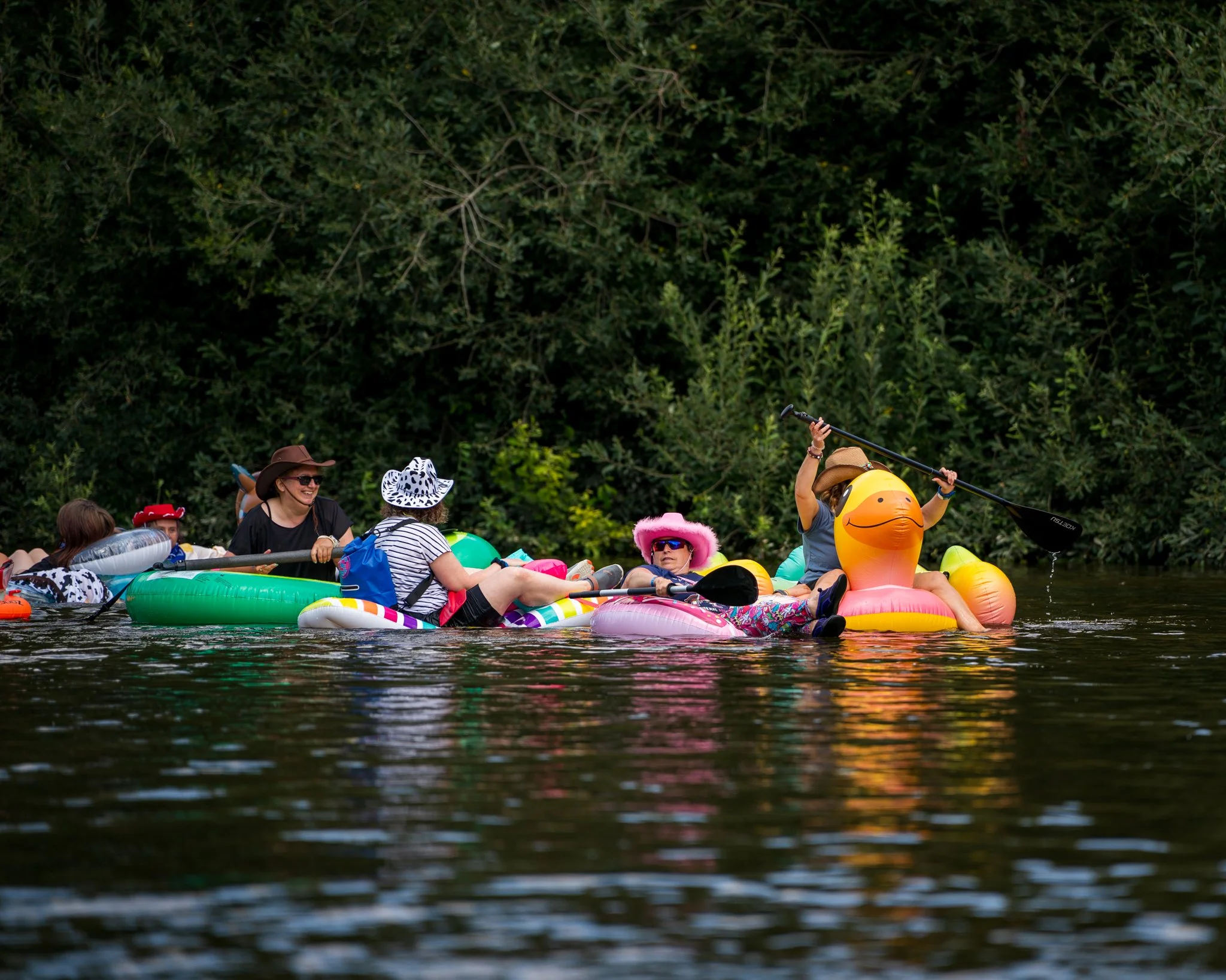 People enjoying a day on a river, sitting on inflatable floats, some wearing hats and sunglasses, with dense green trees in the background.
