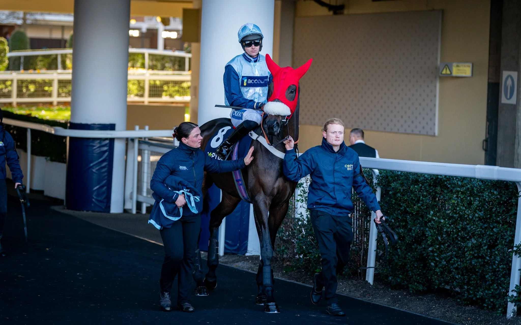 A jockey riding a horse at the racetrack, wearing a red hood with horns, surrounded by handlers.