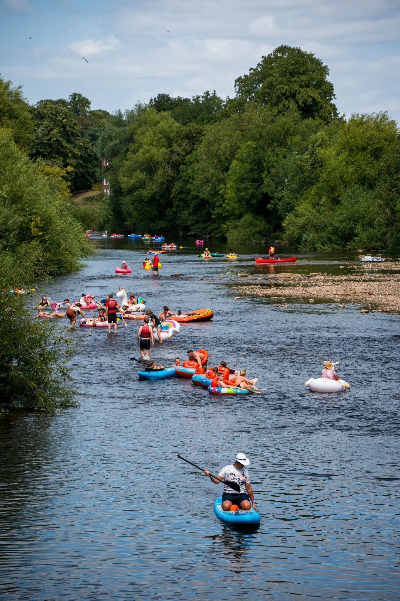 People enjoying a sunny day at a river, floating on colorful inflatable boats and kayaks, surrounded by lush green trees.