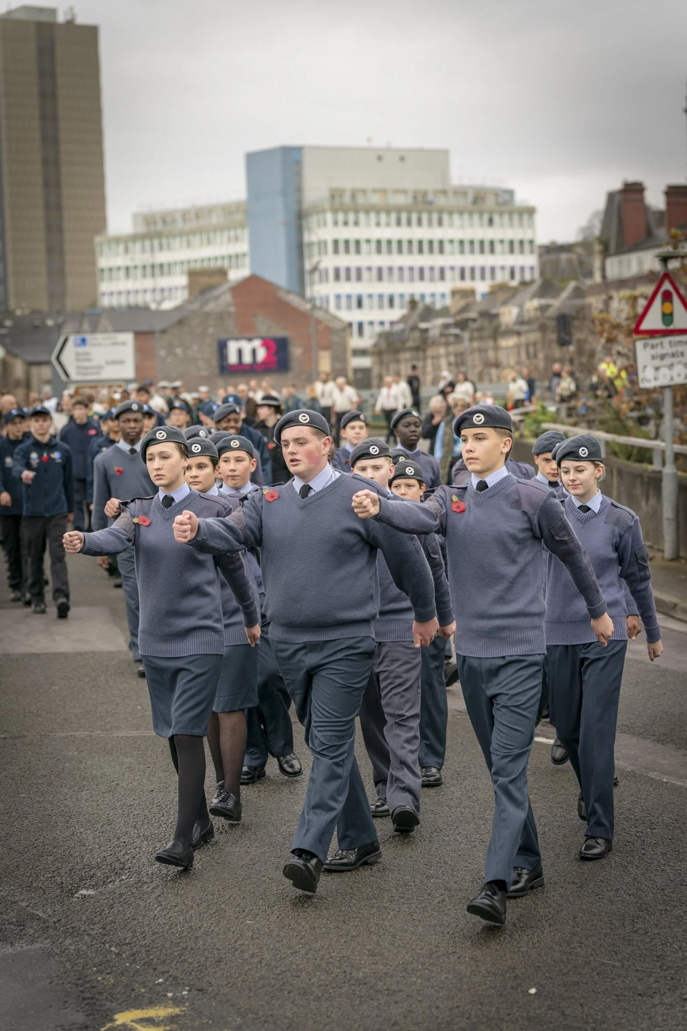 A group of young cadets in uniform marching in a parade, with some wearing poppy pins on their sweaters, against an urban backdrop with buildings and cloudy sky.