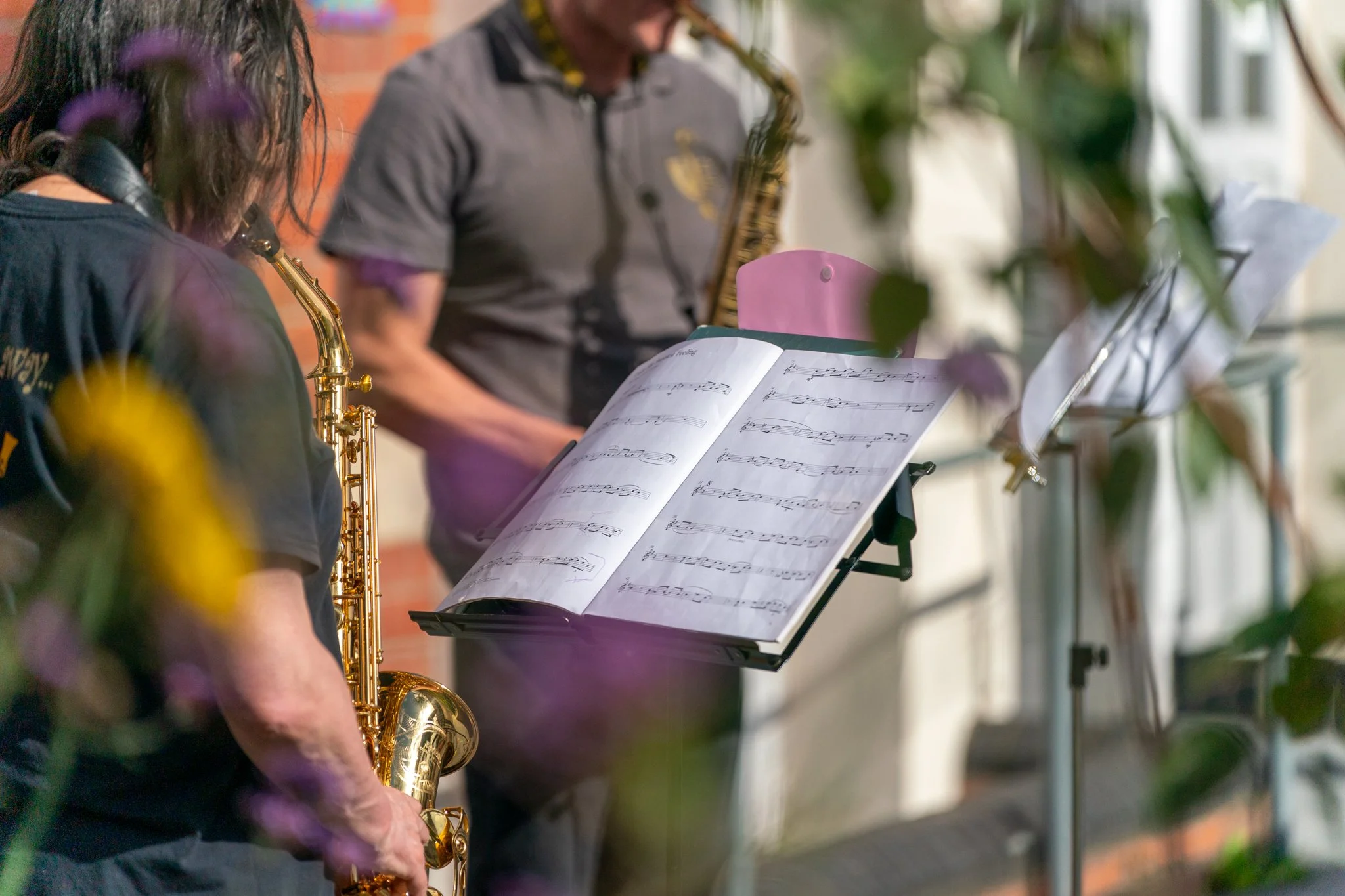 Musicians playing saxophones and reading sheet music outdoors, with flowers partially obscuring the view.