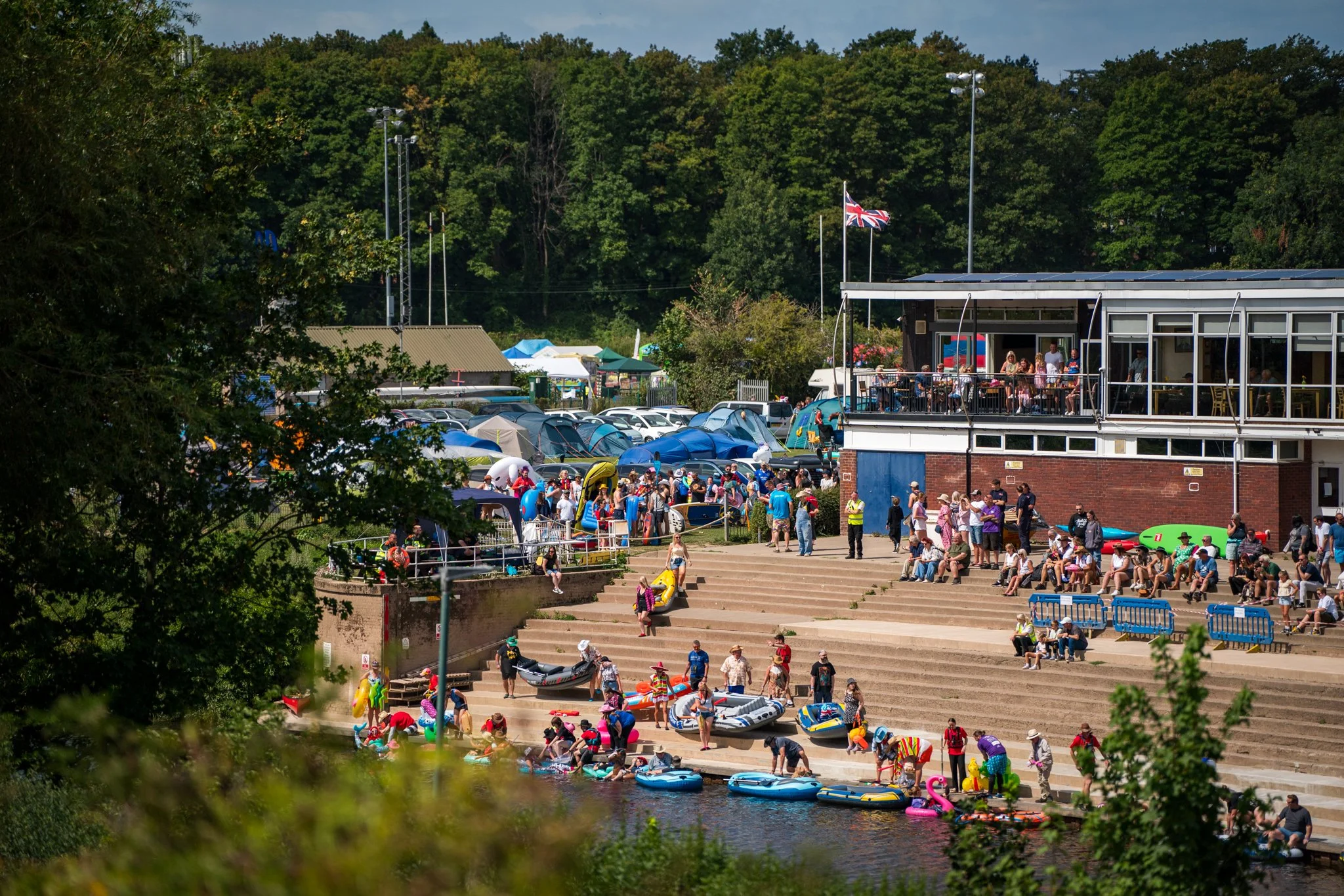 Crowd of people enjoying a sunny day by a river with kayaks and inflatable boats, surrounded by greenery and a building with a balcony flying the Union Jack.