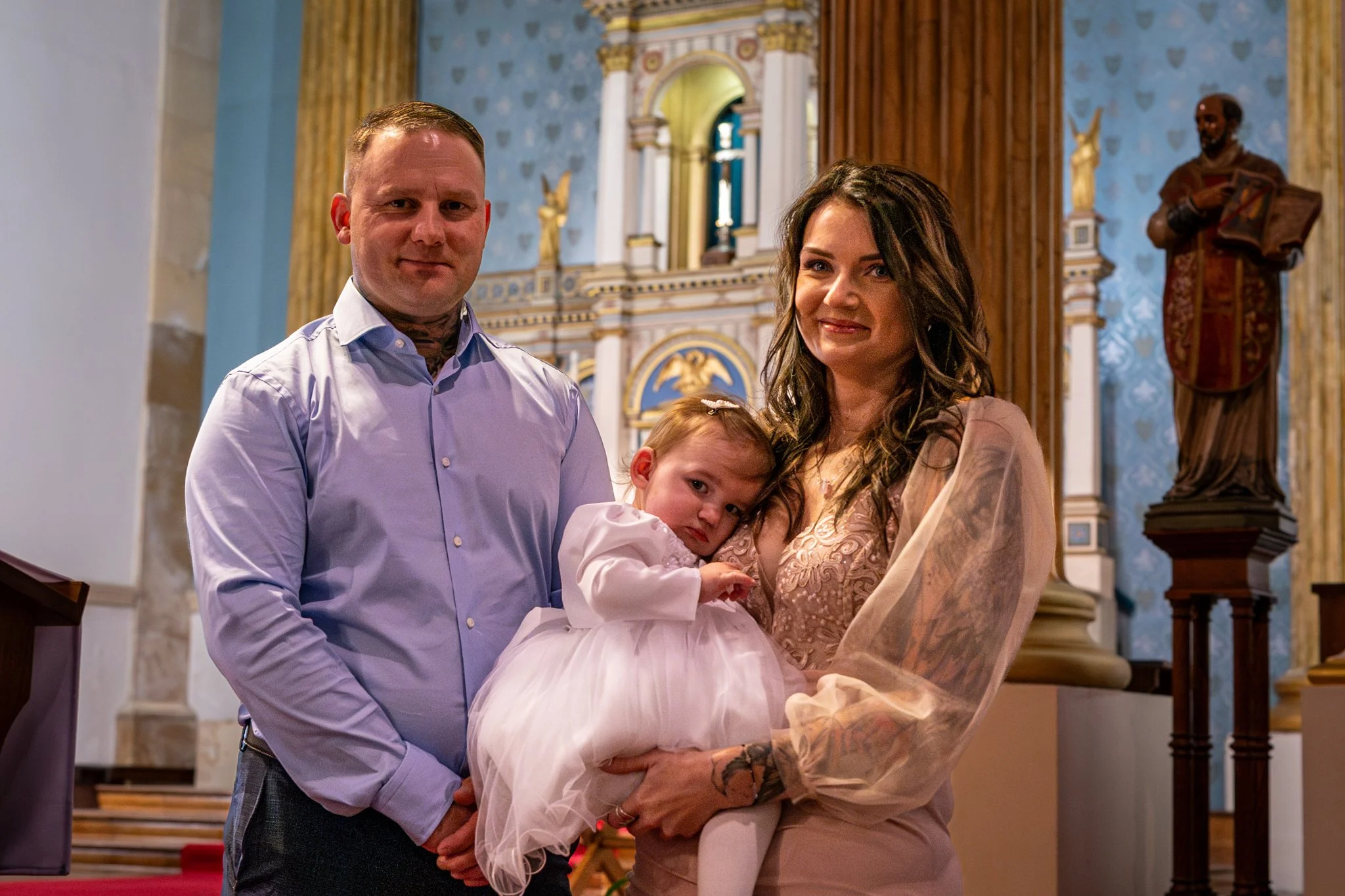 A family of three standing inside a church during a religious service. The man is wearing a light purple dress shirt, the woman is dressed in a pink lace dress, and the young girl is in a white dress with pink accents. The woman holds the girl as the