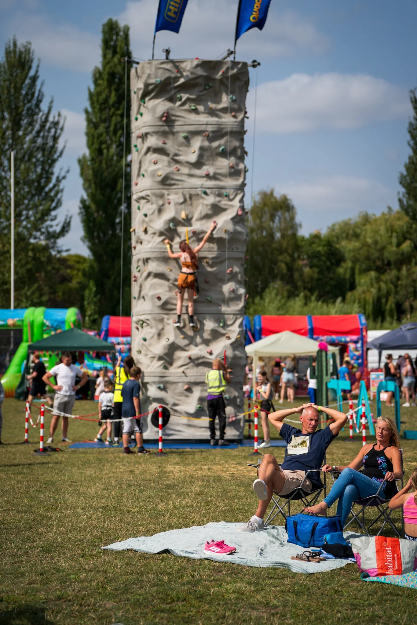 A woman climbing a rock wall at an outdoor festival with flags and tents in the background. People are sitting and standing on the grass, some watching and others walking around.