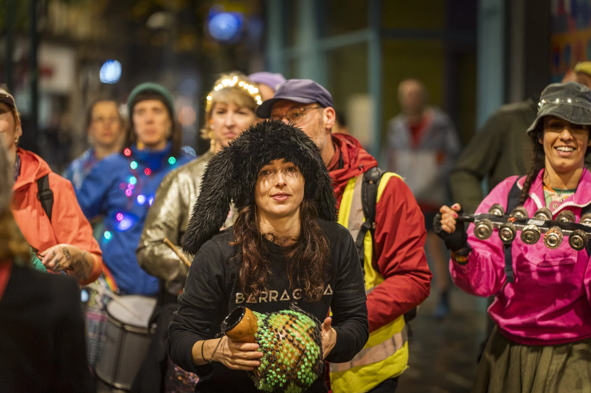 Group of people celebrating on a city street at night, some wearing costumes and accessories, with festive decorations, including string lights, and people smiling and dancing.