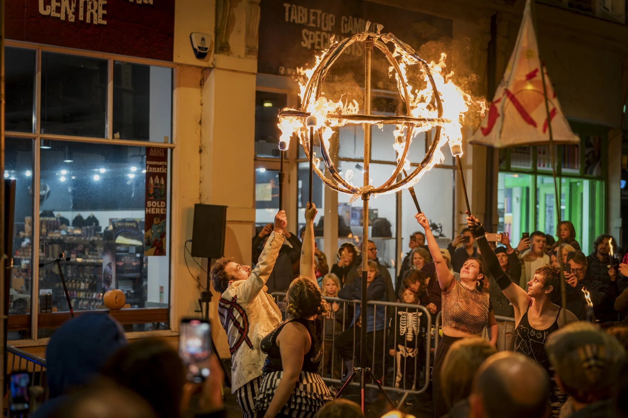 People gather around and watch a fire demonstration where individuals are holding long sticks with a flaming globe structure made of fire, inside a venue with spectators and some taking photos.
