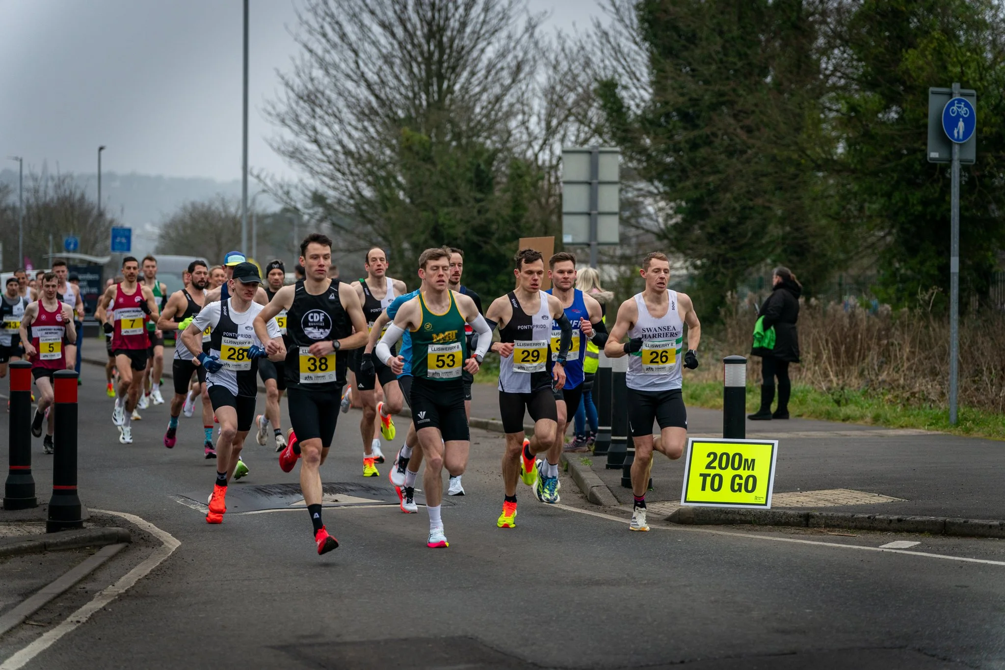 A group of marathon runners running on a street during a race. There are some signs on the street, including a yellow sign that reads '200M TO GO'. It is overcast and trees line the street.