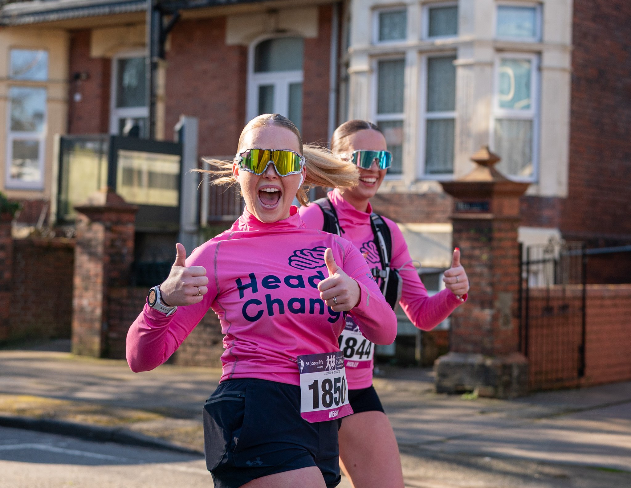 Two women running in a road race, both wearing pink shirts, sunglasses, and race bibs with numbers.